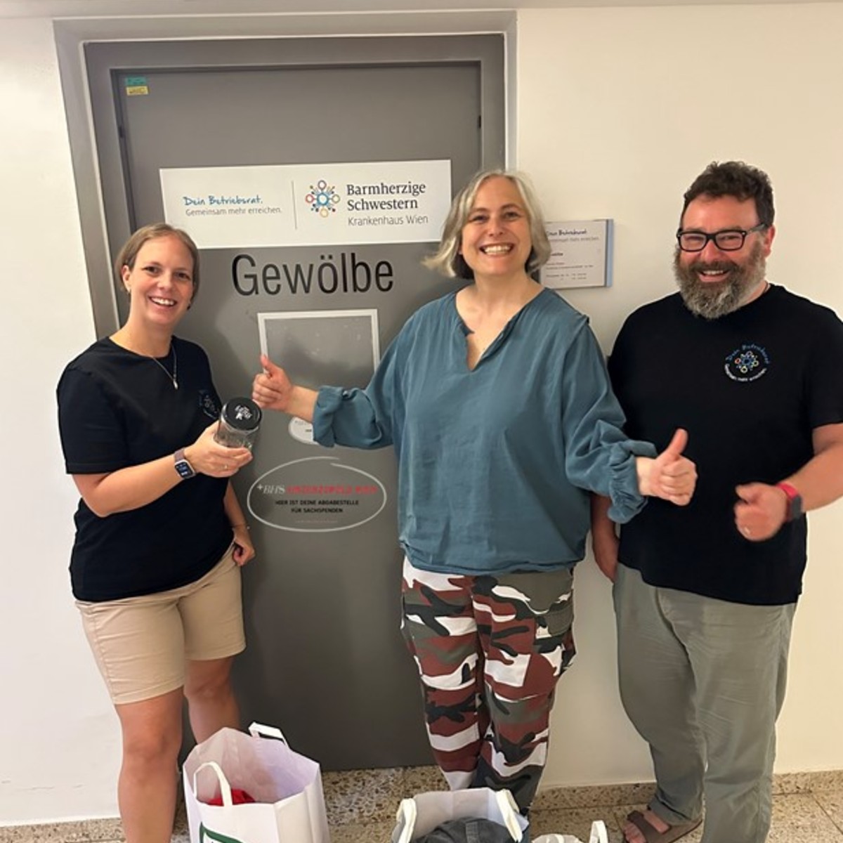 Zwei Frauen und ein Mann mit Bart vor einer Türe, auf der in großen Lettern "Gewölbe" steht. Am Boden vor ihnen sind Einkaufstaschen mit Spenden.