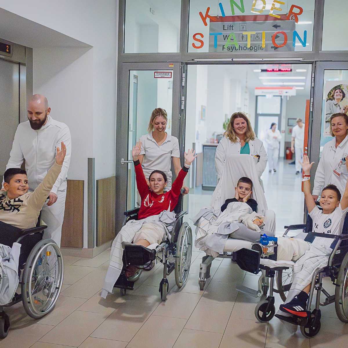 Ein Gang in der Kinderstation. Vier Kinder werden in Rollstühlen von Pflegekräften und Mediziner*innen transportiert. Einige haben Verbände an den Beinen, drei der Kinder strecken jubelnd die Hände in die Höhe.