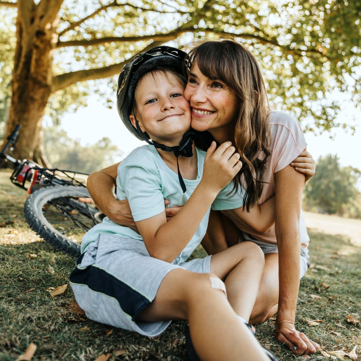 Eine Mutter mit Sohn im Freien unter einem Baum, hinter dem Kind liegt ein Fahrrad, am Knie hat er ein Pflaster. Die Mutter umarmt den Jungen, beide lächeln.