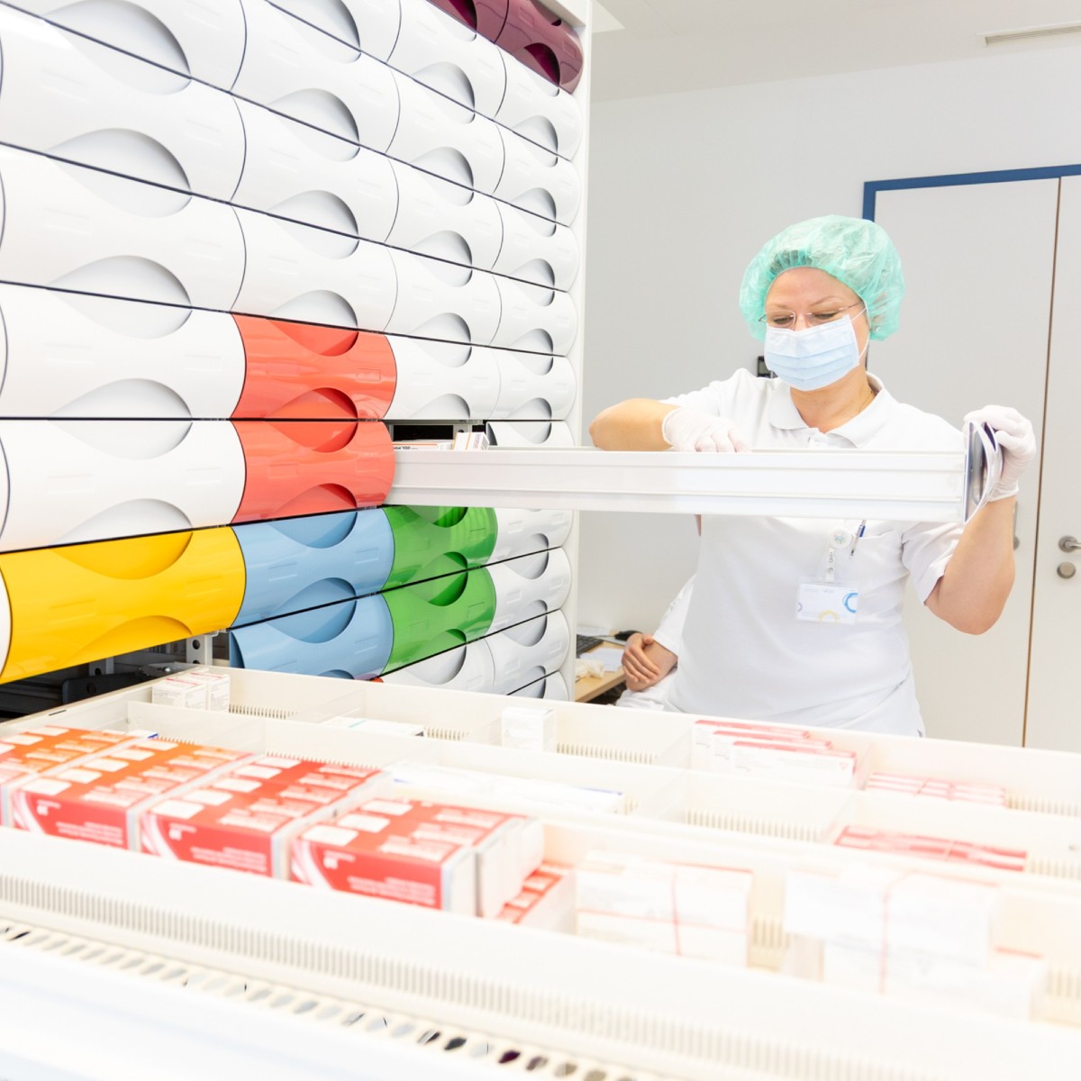 Eine Mitarbeiterin in einer Apotheke in Dienstkleidung mit Maske und Haarnetz. Sie hat eine von vielen Schubladen eines Medikamentenarchivs geöffnet und durchsucht die Lade.