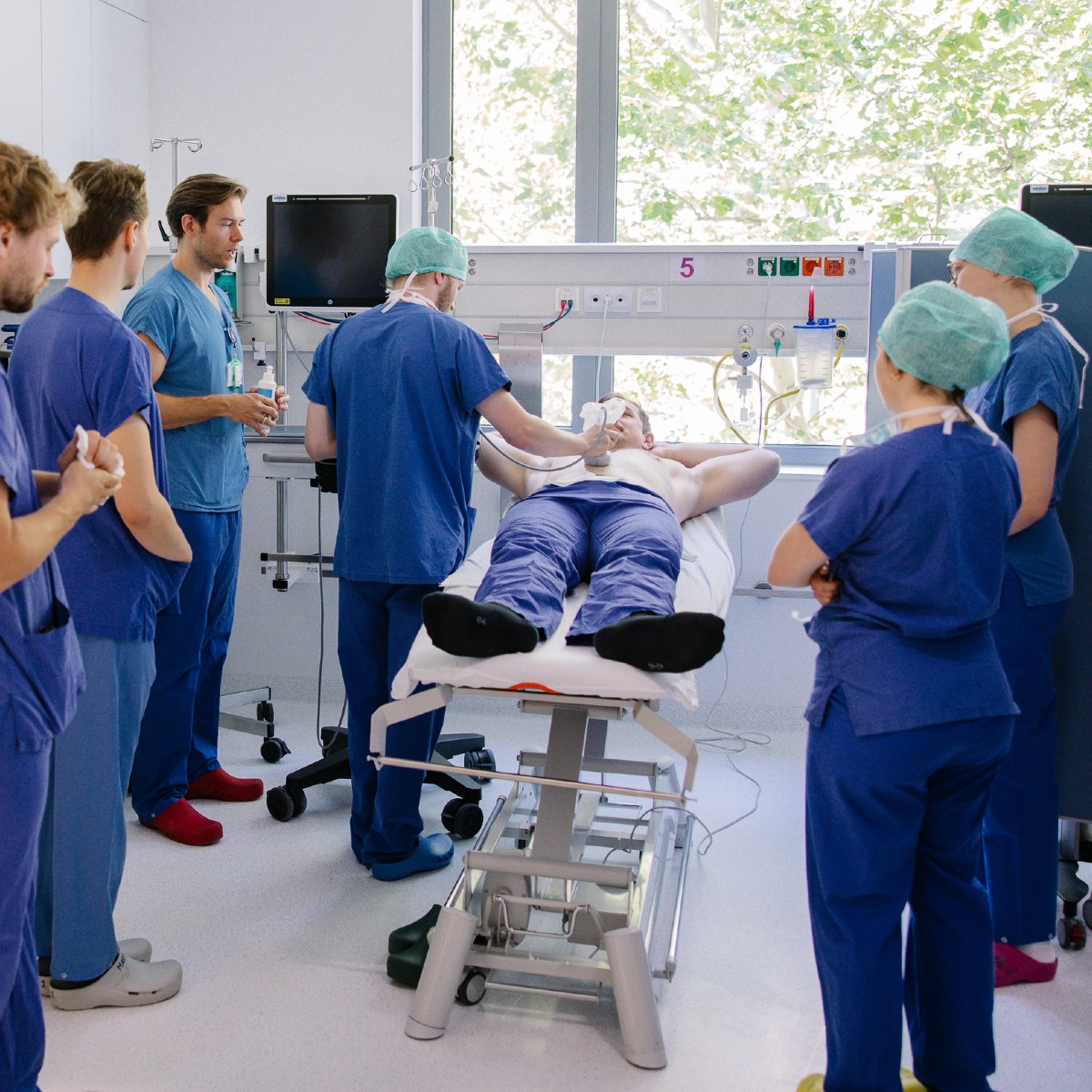 Einige junge angehende Mediziner*innen in blauer Arbeitskleidung stehen um einer Liege. Darauf liegt ein Schauspiel-Patient mit unbekleidetem Oberkörper, einer der angehenden Ärzte führt einen Ultraschall durch.