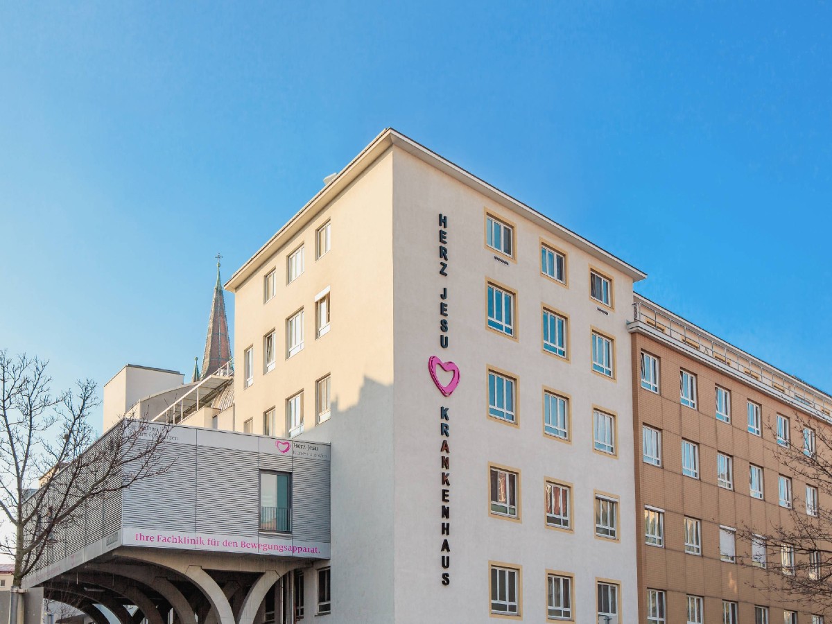 Eine Außenaufnahme des Herz-Jesu Krankenhauses. Das Gebäude ist eingebettet zwischen Bäumen, der Himmel darüber strahlend blau. Markant ist der senkrechte Schriftzug HERZ JESU KRANKENHAUS mit einem Herz in der Mitte.