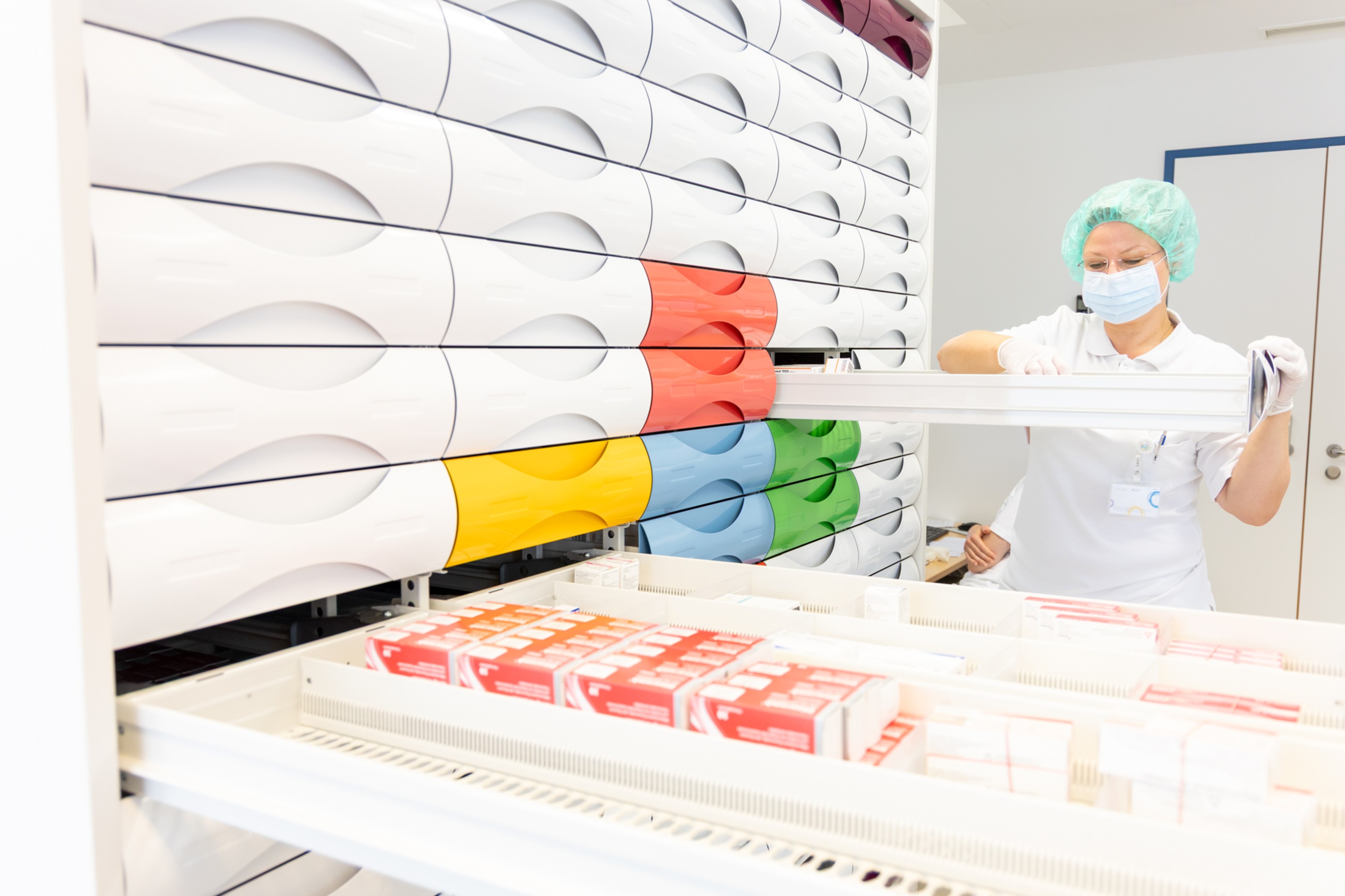 Eine Mitarbeiterin in einer Apotheke in Dienstkleidung mit Maske und Haarnetz. Sie hat eine von vielen Schubladen eines Medikamentenarchivs geöffnet und durchsucht die Lade.