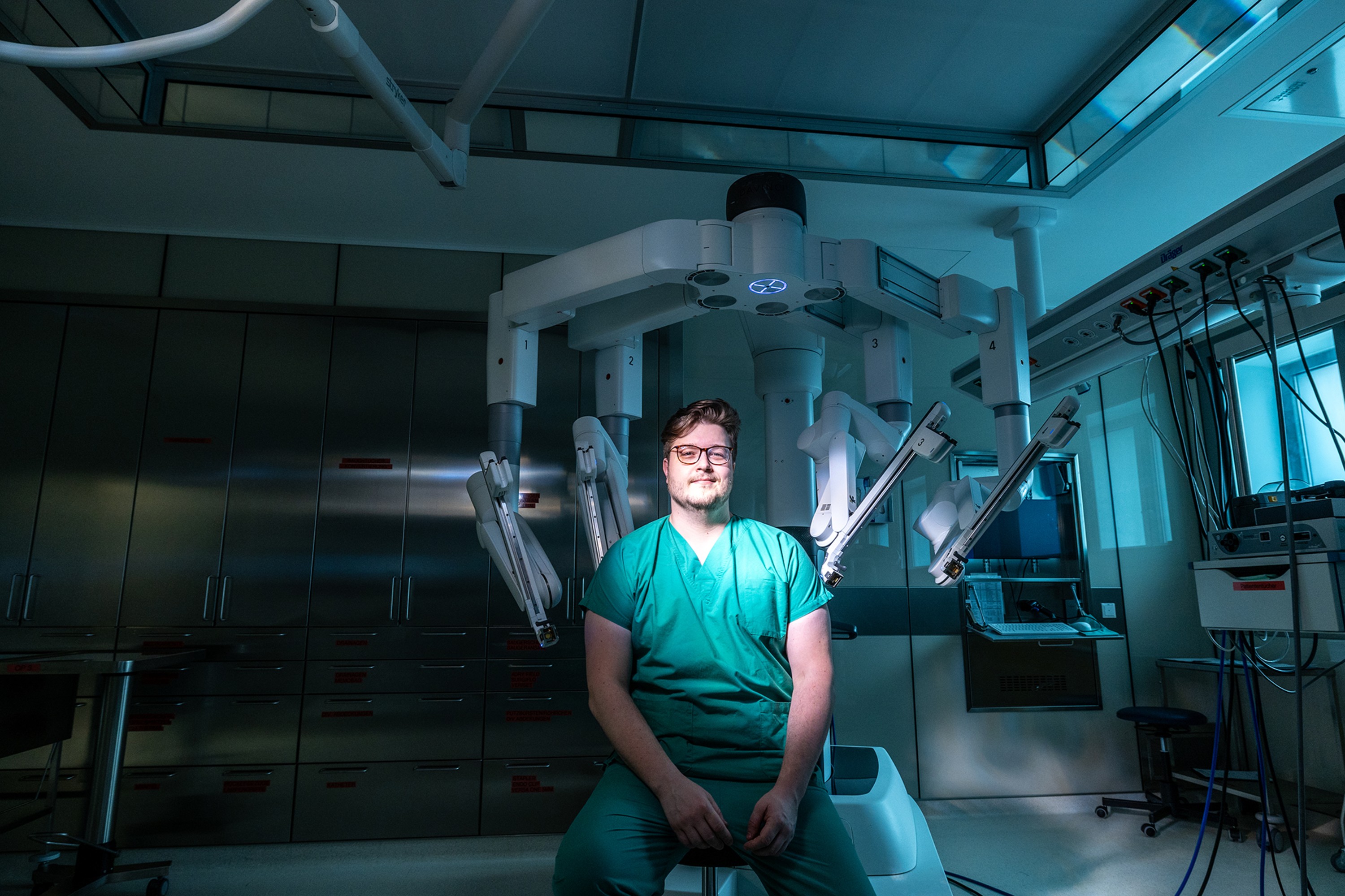 Ein Arzt sitzt vor einem OP-Roboter in einem dunklen Operationssaal. Nur der Arzt und Teile des Roboters sind beleuchtet. Der Arzt lächelt.