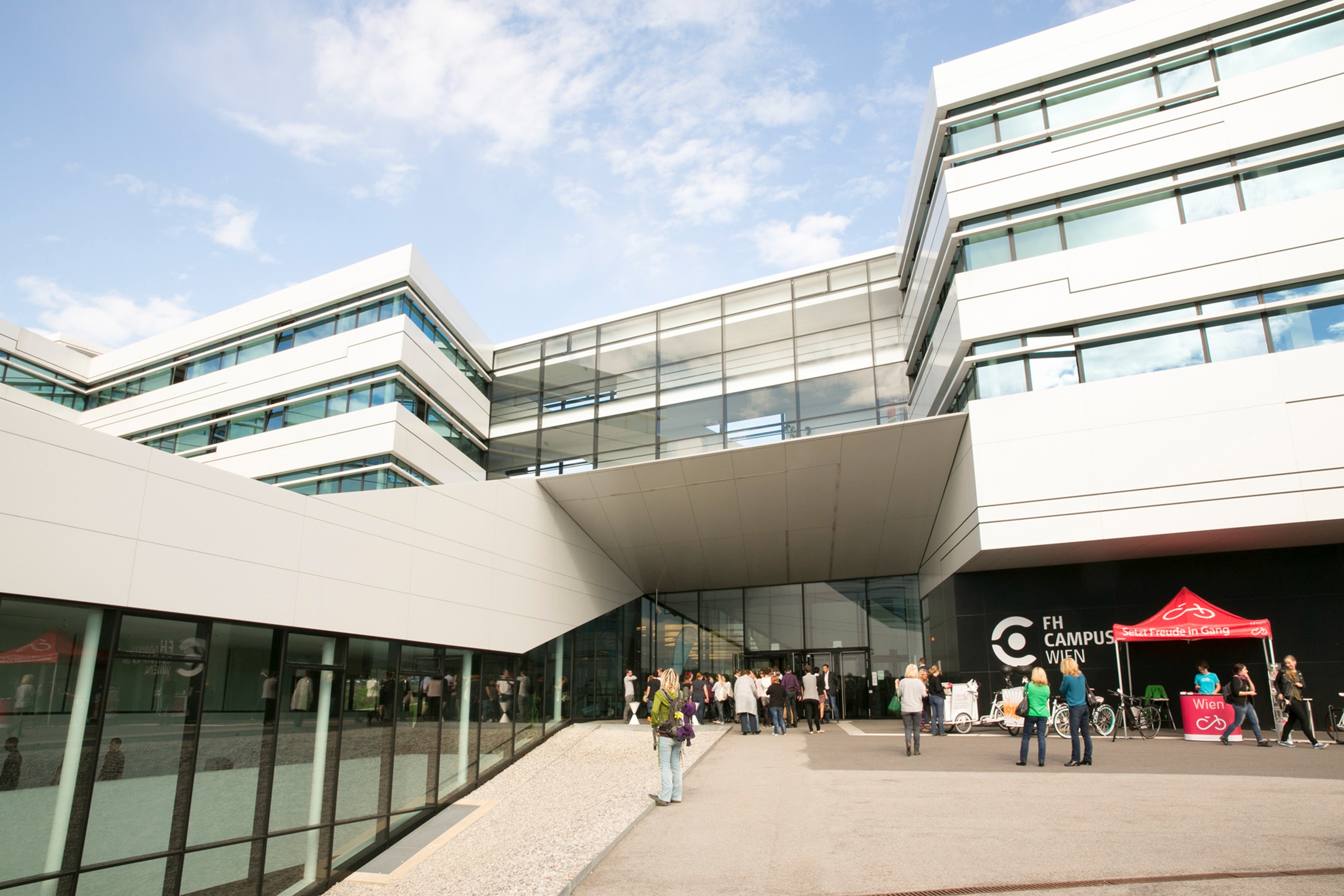 Eine Außenansicht des FH Campus Wien. Ein modernes, kantiges Gebäude. Beim Eingang tummeln sich zahlreiche Menschen.