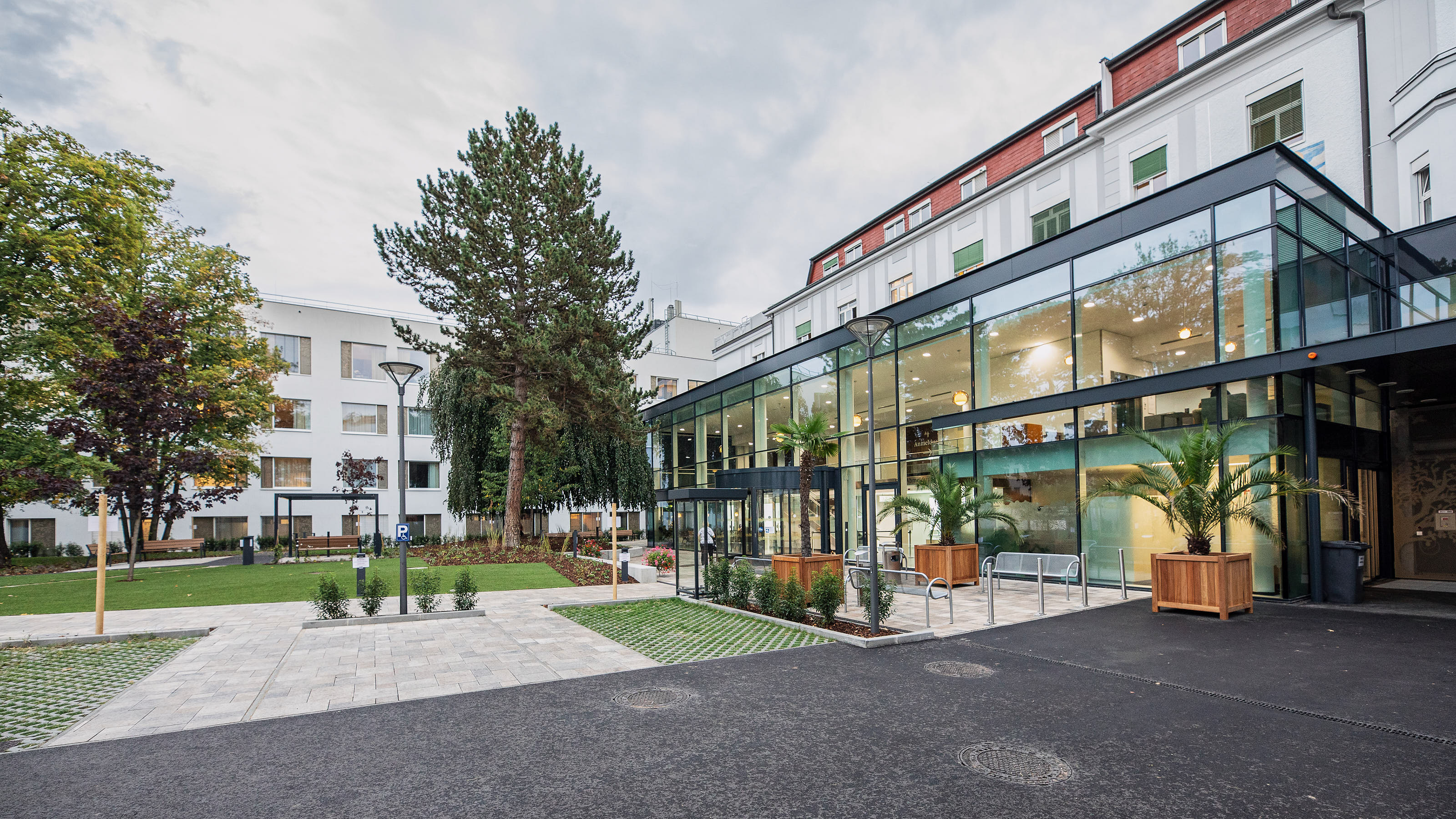 Eine Außenaufnahme des St. Josef Krankenhauses Wien, auf der der Eingangsbereich zu sehen ist. Er ist mit viel Glas gestaltet, Bäume und kleine Palmen schmücken den Weg aus