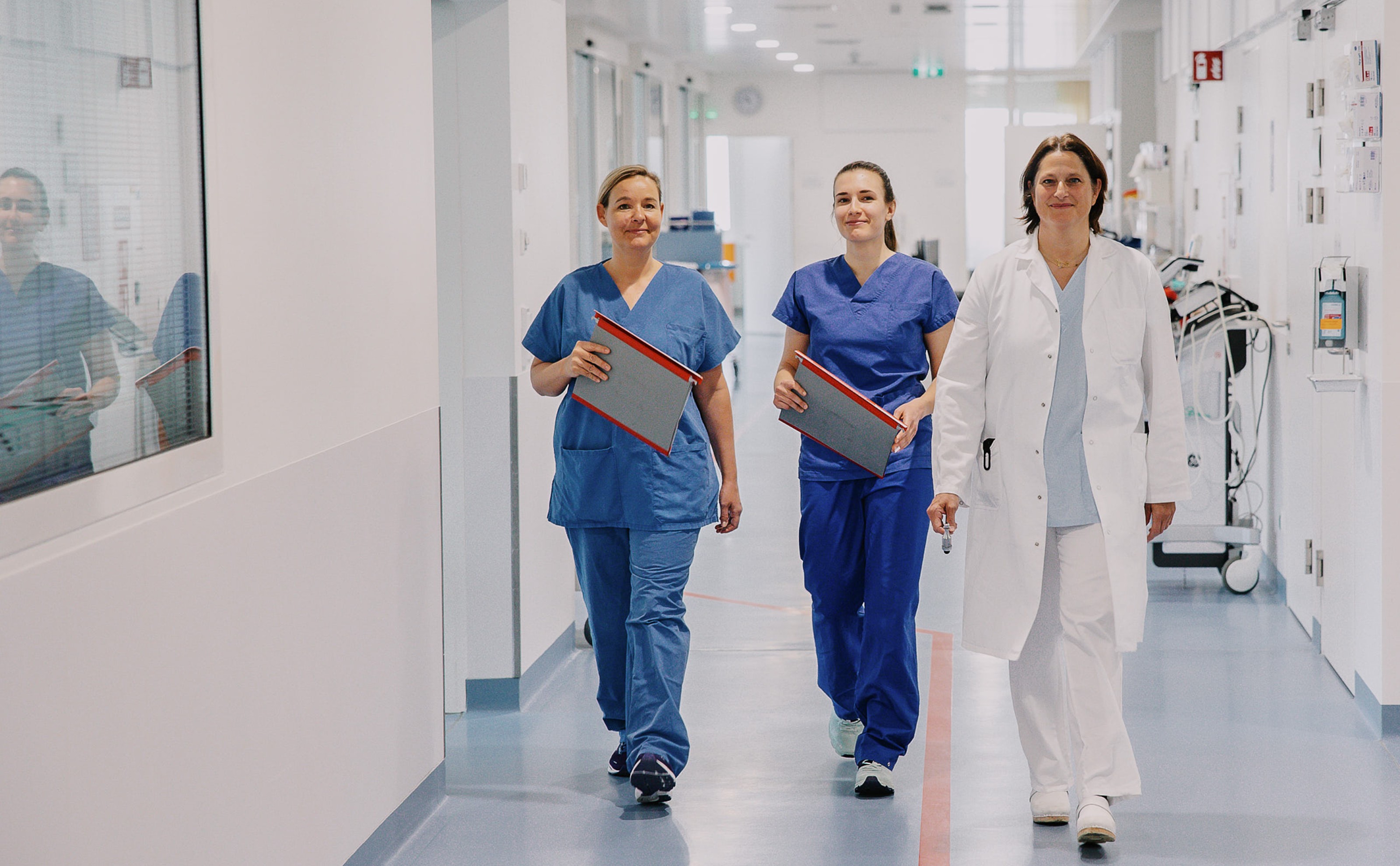 Eine Ärztin im weißen Kittel und zwei Frauen in Krankenhauskleidung gehen einen Gang im Krankenhaus entlang.