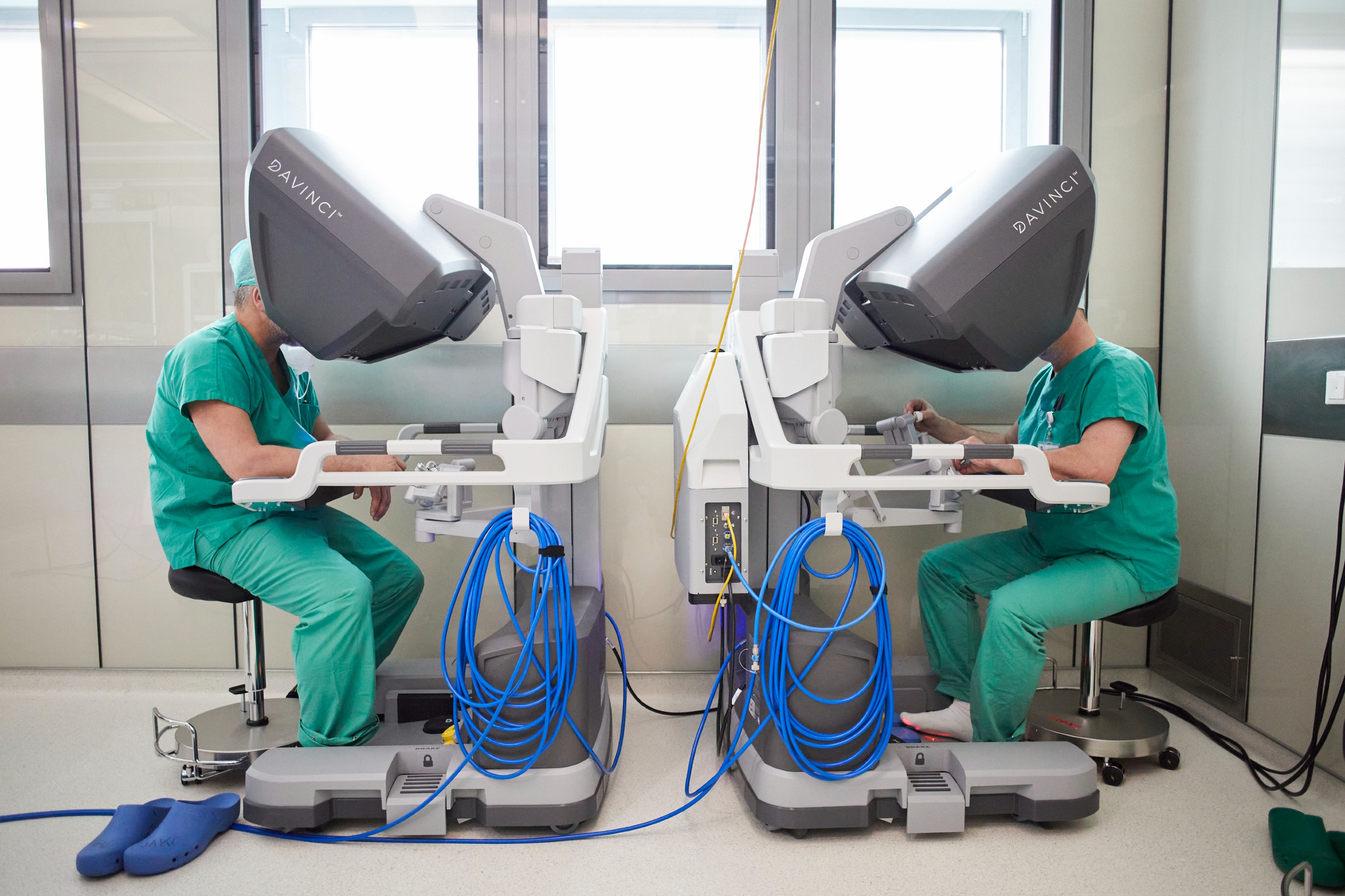 Zwei Mediziner*innen sitzen gegenüber, jeder steuert einen Davinci-OP-Roboter. 