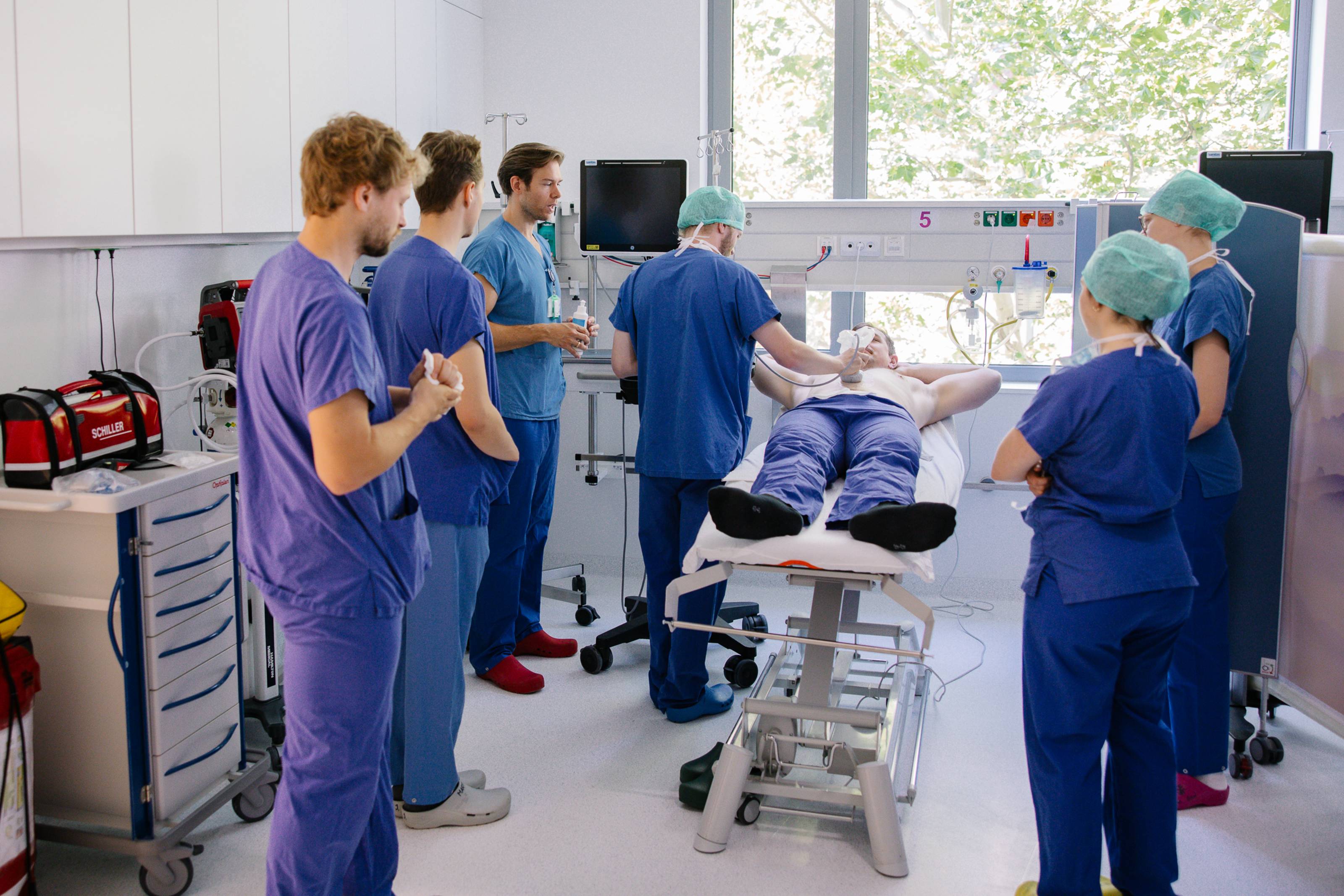 Einige junge angehende Mediziner*innen in blauer Arbeitskleidung stehen um einer Liege. Darauf liegt ein Schauspiel-Patient mit unbekleidetem Oberkörper, einer der angehenden Ärzte führt einen Ultraschall durch.