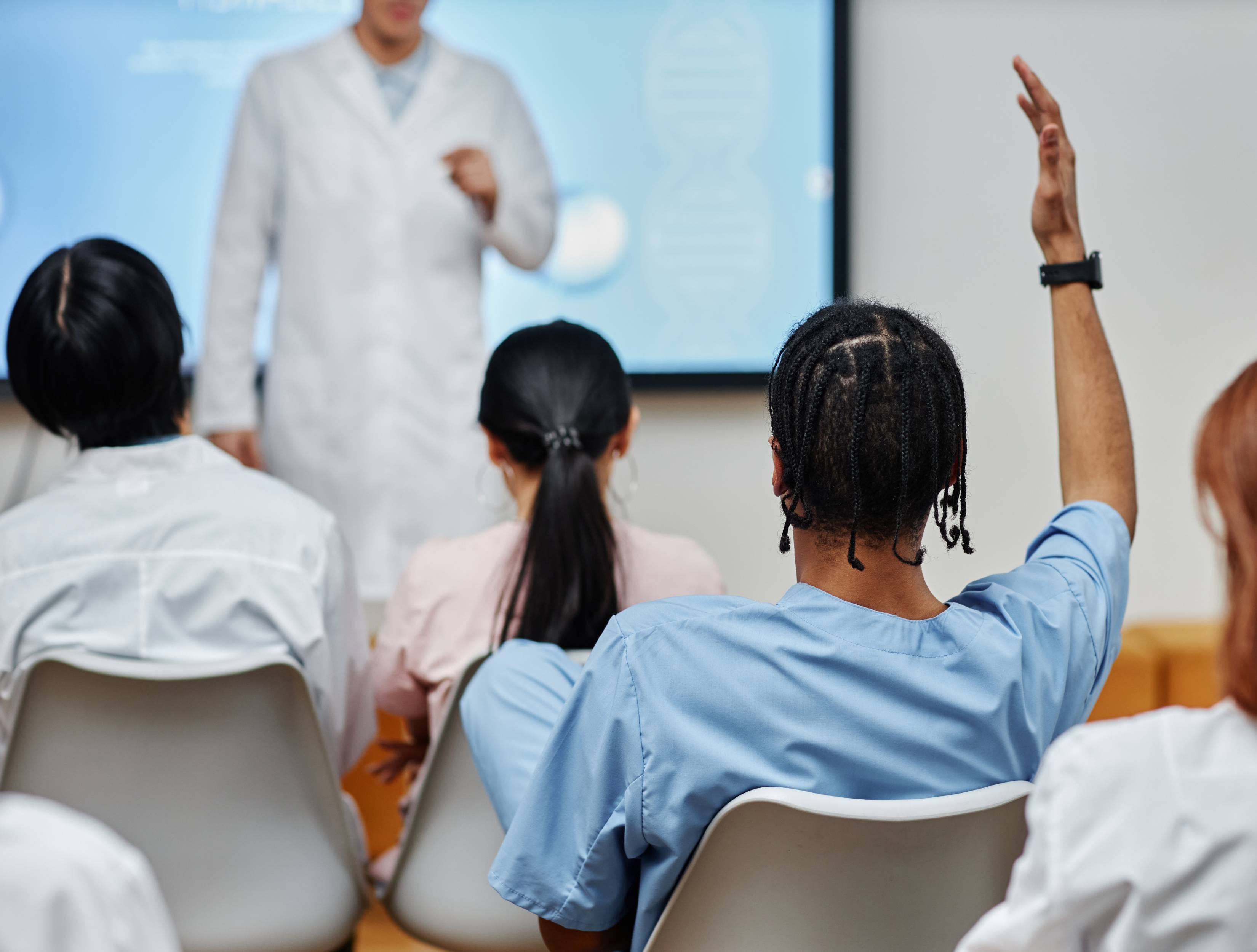Rückansicht von einem jungen Medizinstudenten mit Dreadlocks, der in einem Schulungsraum aufzeigt. Im Hintergrund verschwommen eine Lehrkraft und andere Schüler*innen.