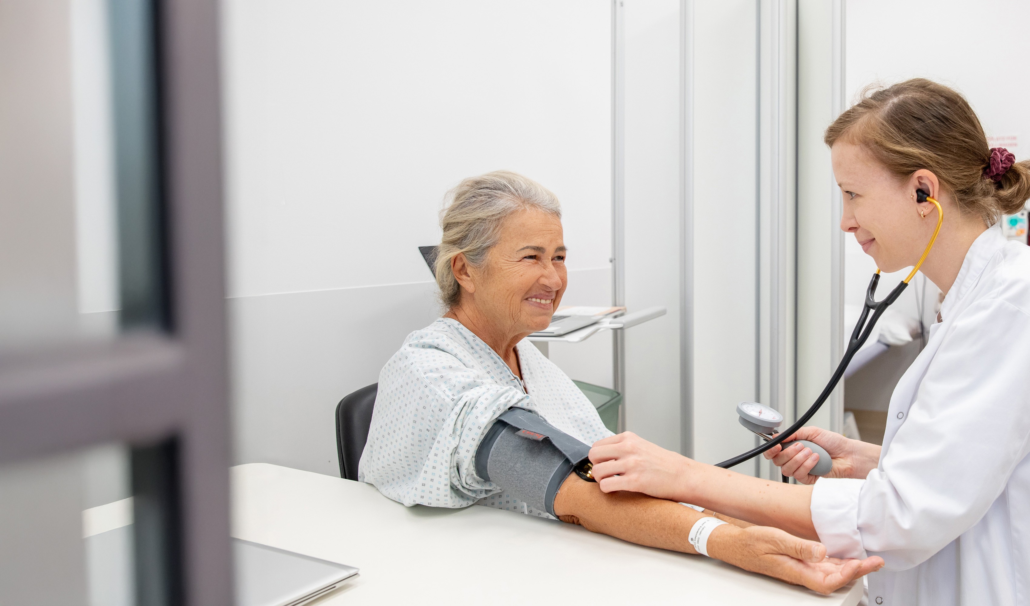 Eine Patientin in einem Behandlungsraum, bei der eine Ärztin im weißen Kittel gerade eine Blutdruckmessung durchführt. Beide sind in einem freundlichen Gespräch vertieft.