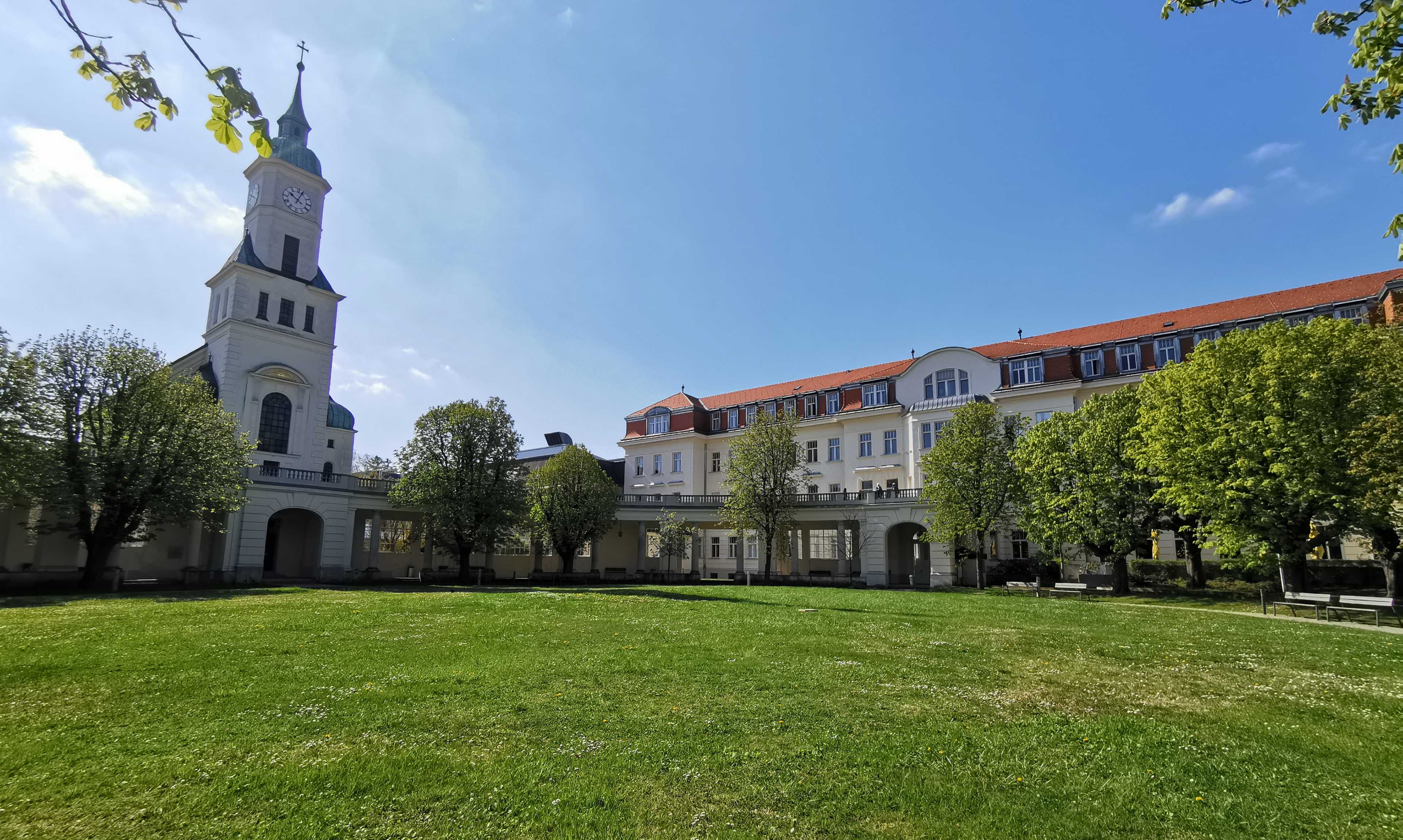 Blick auf das Orthopädische Spital Speising. Vor der Klinik ist ein Park mit einer großen Grünfläche mit Bäumen, links vom Gebäude eine Kirche 