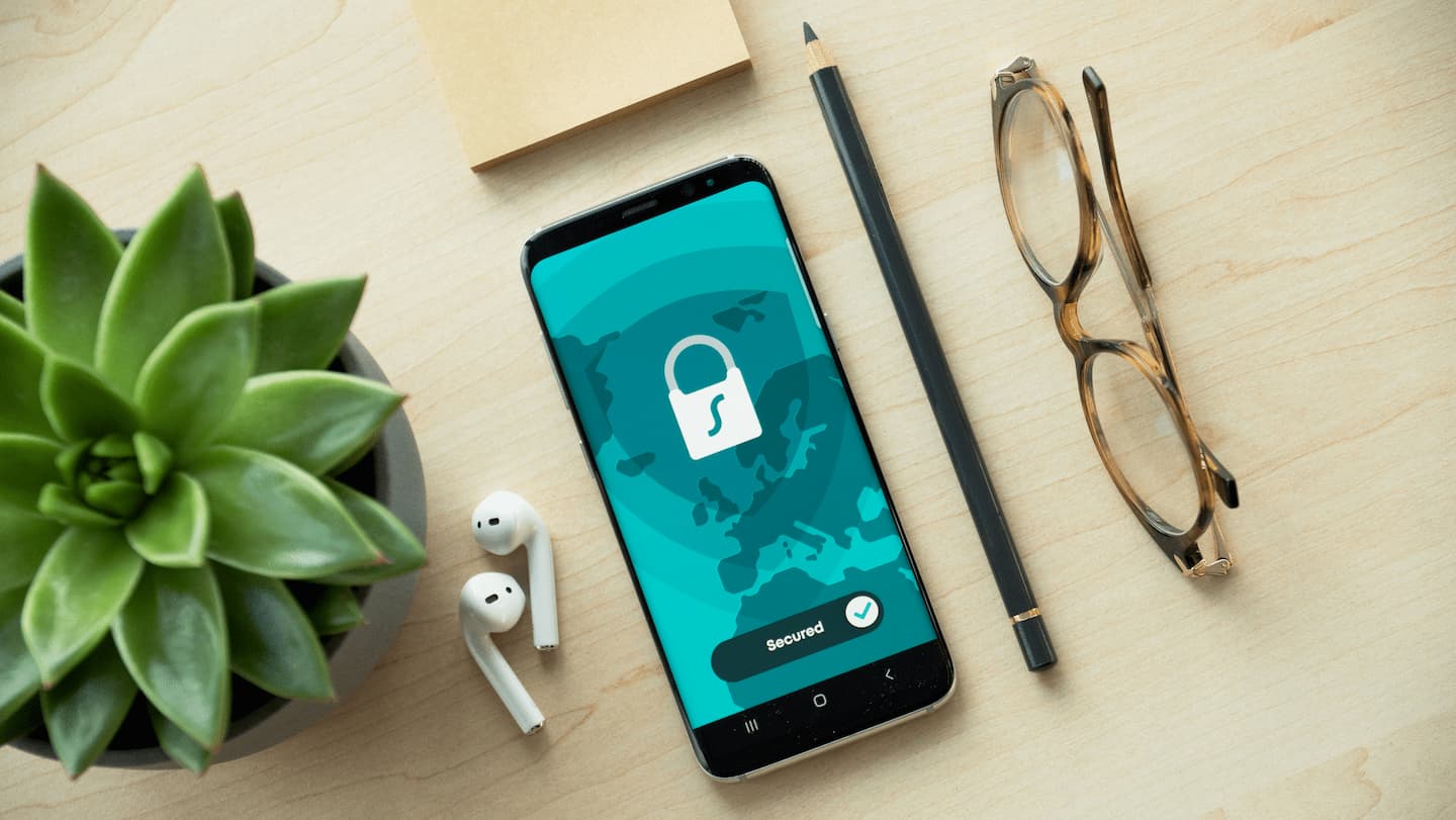 Smartphone with a lock icon showing secured status, next to a plant, pencil, glasses, earbuds, and notepad on a wooden surface.