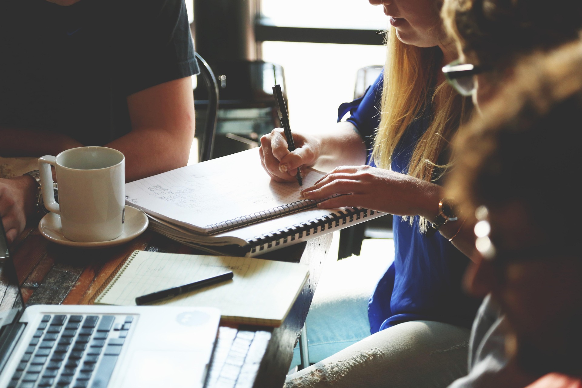 People collaborating around a table with notebooks, a laptop, and a coffee cup, focused on writing and discussing.