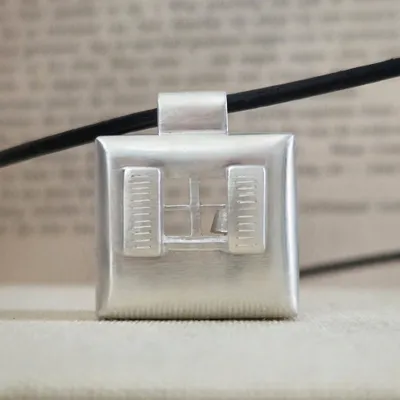 Silver pendant shaped like a window with shutters, featuring a small sailboat inside. Background is blurred text.