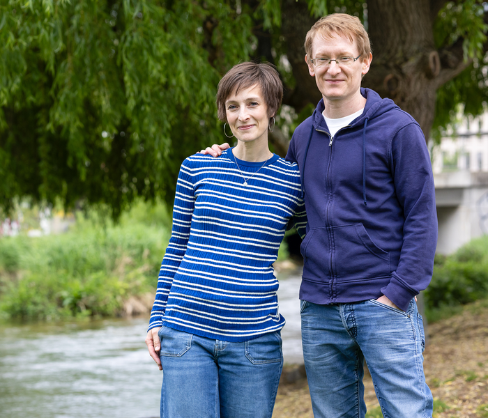 Ein lächelndes Paar steht an einem Flussufer, mit üppigem Grün und einem Baum im Hintergrund. Die Frau trägt einen blau gestreiften Pullover, der Mann einen blauen Kapuzenpulli.