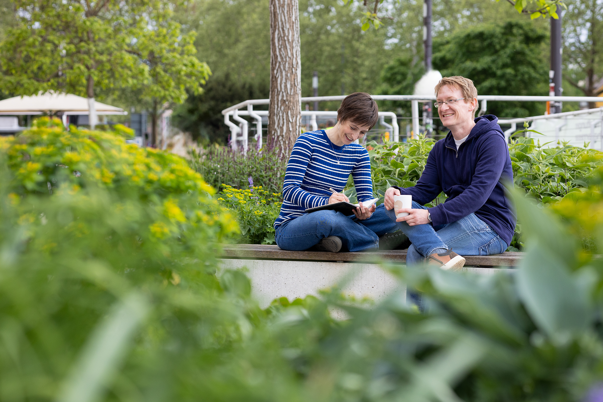 Zwei Menschen sitzen auf einer Bank in einem üppigen Garten, lächeln und schreiben in Notizbücher, umgeben von Grün und Bäumen.