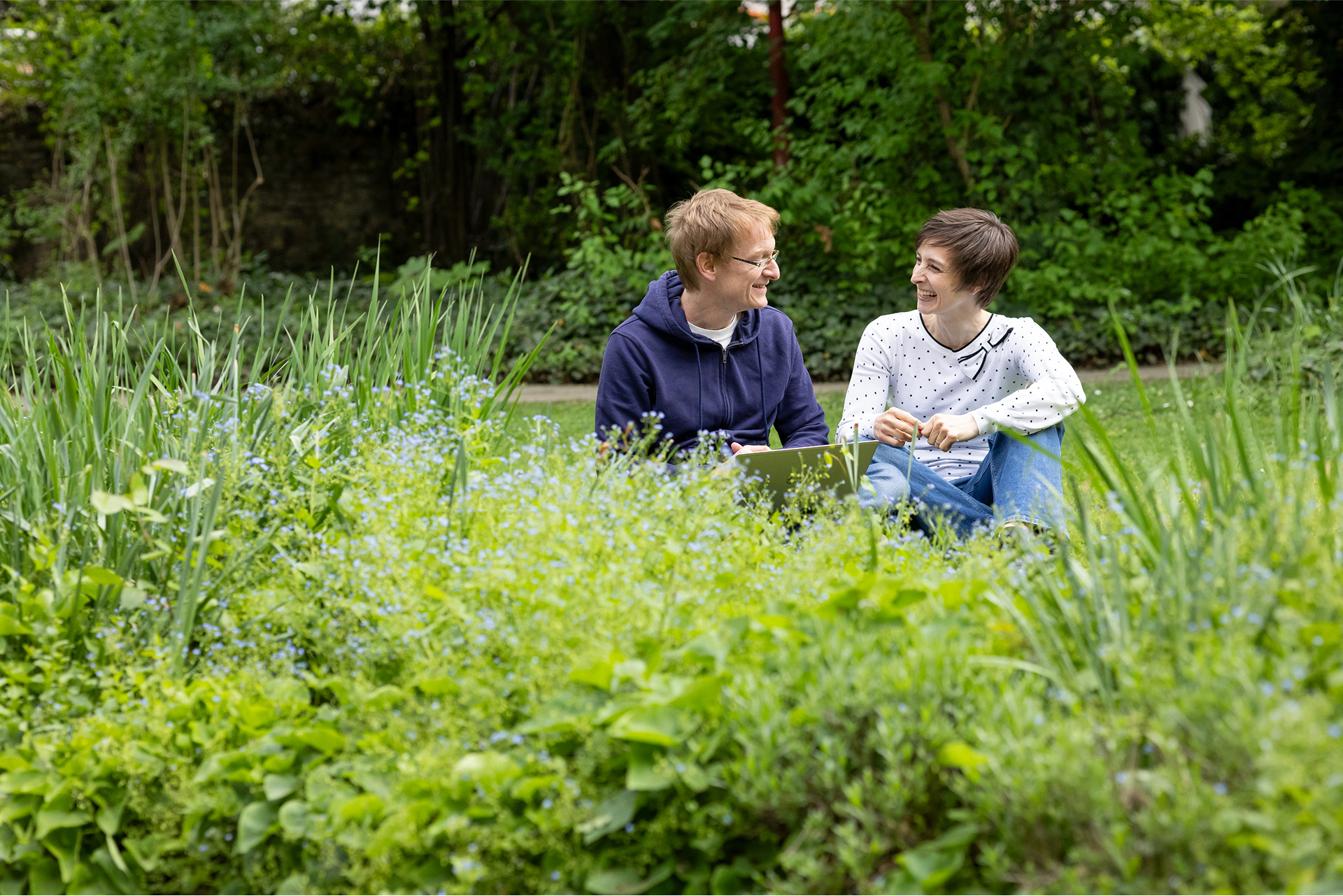 Zwei Menschen, die in einem üppigen Garten im Gras sitzen und sich gegenseitig anlächeln, umgeben von Grün und Blumen.