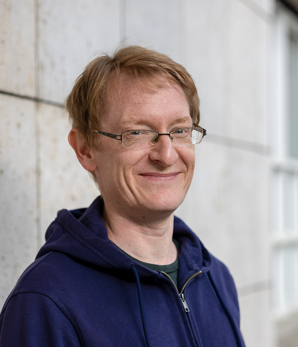 Eine Person mit Brille und blauem Kapuzenpulli steht lächelnd vor einer hellen, strukturierten Wand.