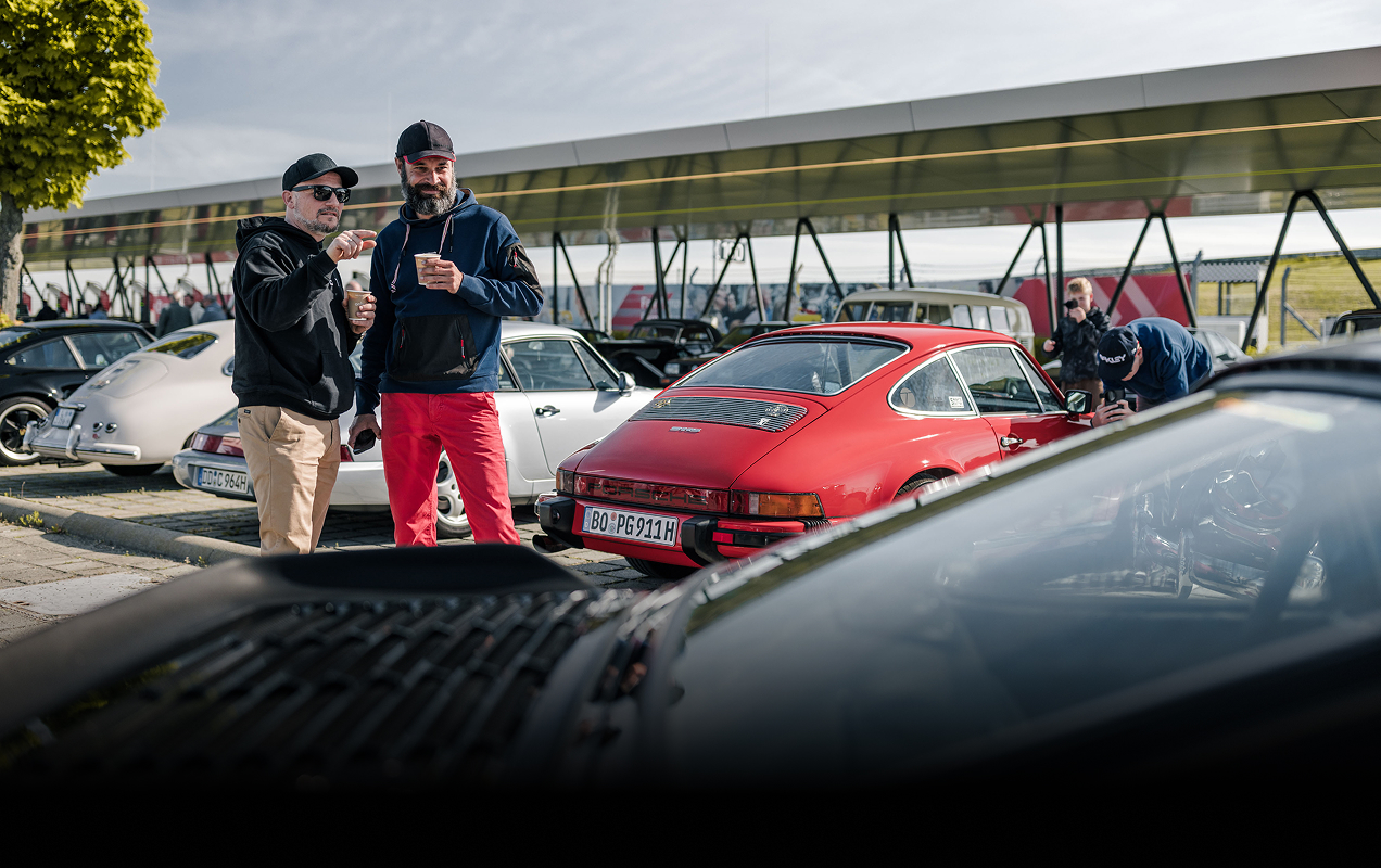 2 Männer mit Kaffeebechern in den Händen betrachten klassische Porsche.