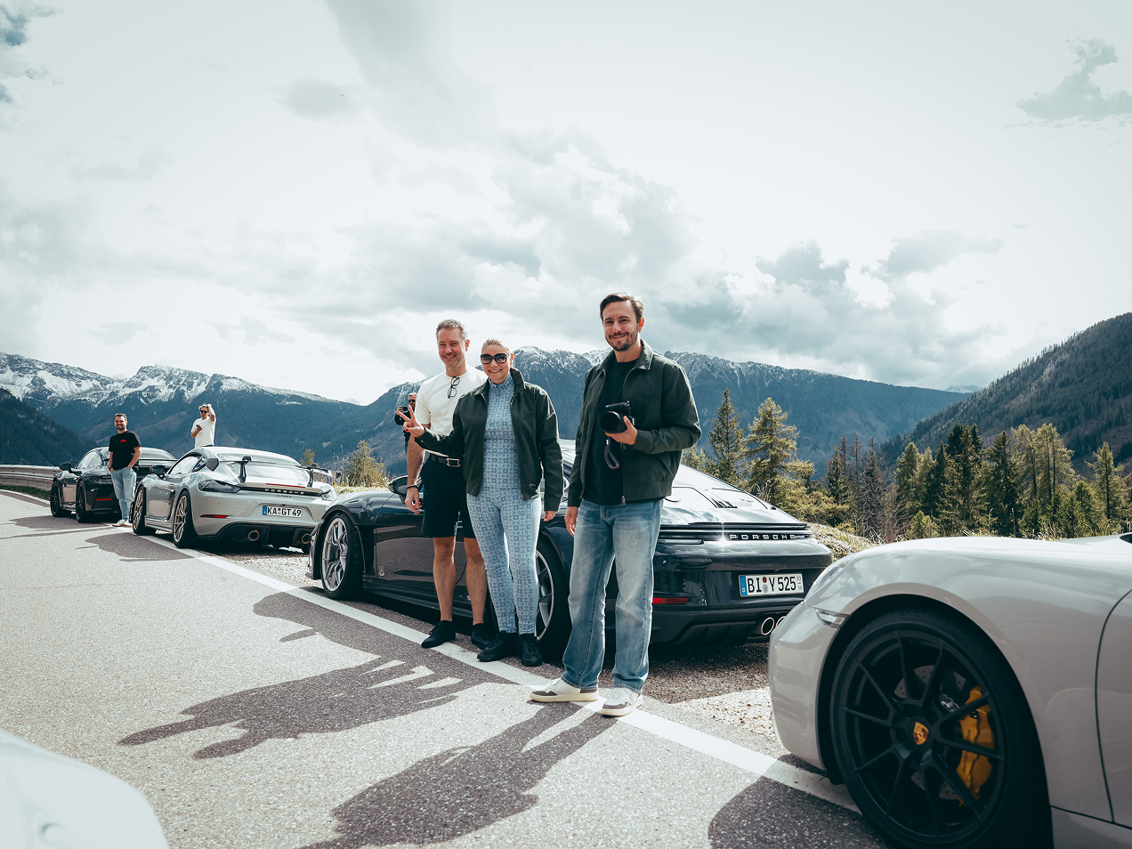 Mehrere Menschen stehen neben ihren Porsche am Straßenrand vor einer Berglandschaft.