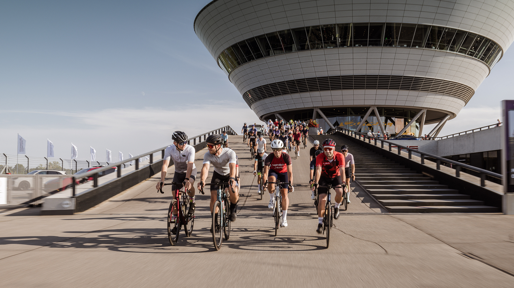 Blick von vorne auf mehrere Rennradfahrer, die die Auffahrt zum Porsche Experience Center Leipzig hinunterfahren.