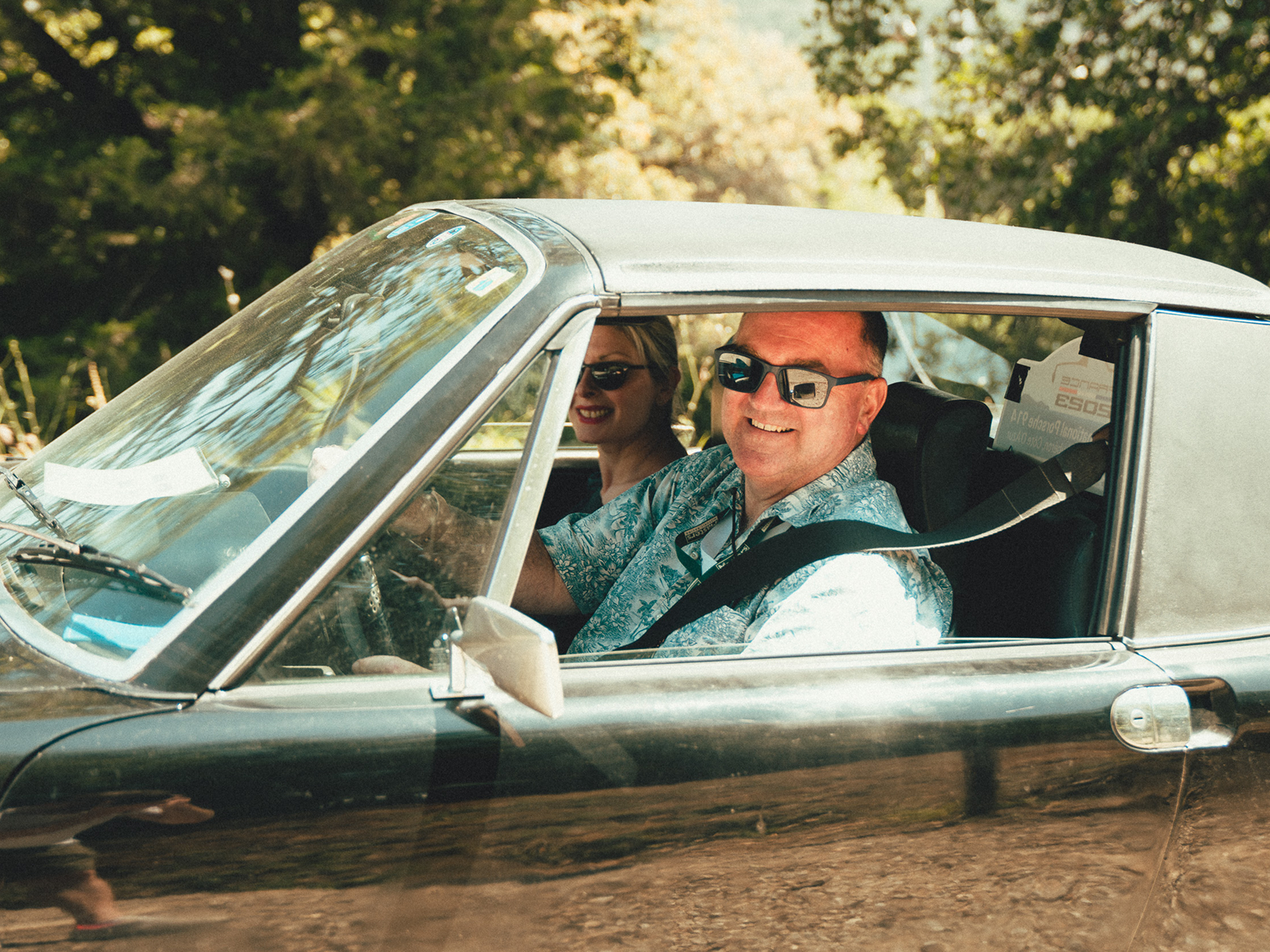 Ein Pärchen mit Sonnenbrille lächelt aus einem Auto in die Kamera.