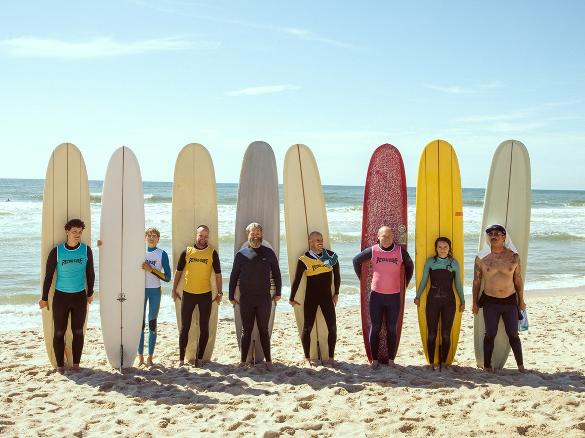 7 Männer und 1 Frau stehen vor ihren Surfbrettern am Strand.