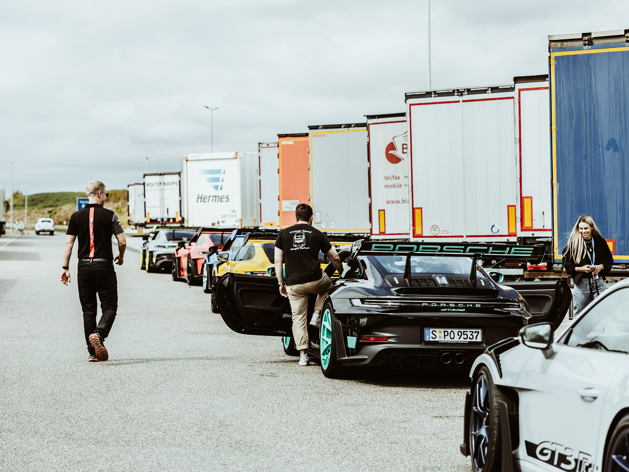 Eine Autokolonne von Porsche Fahrzeugen stehen neben LKW's.