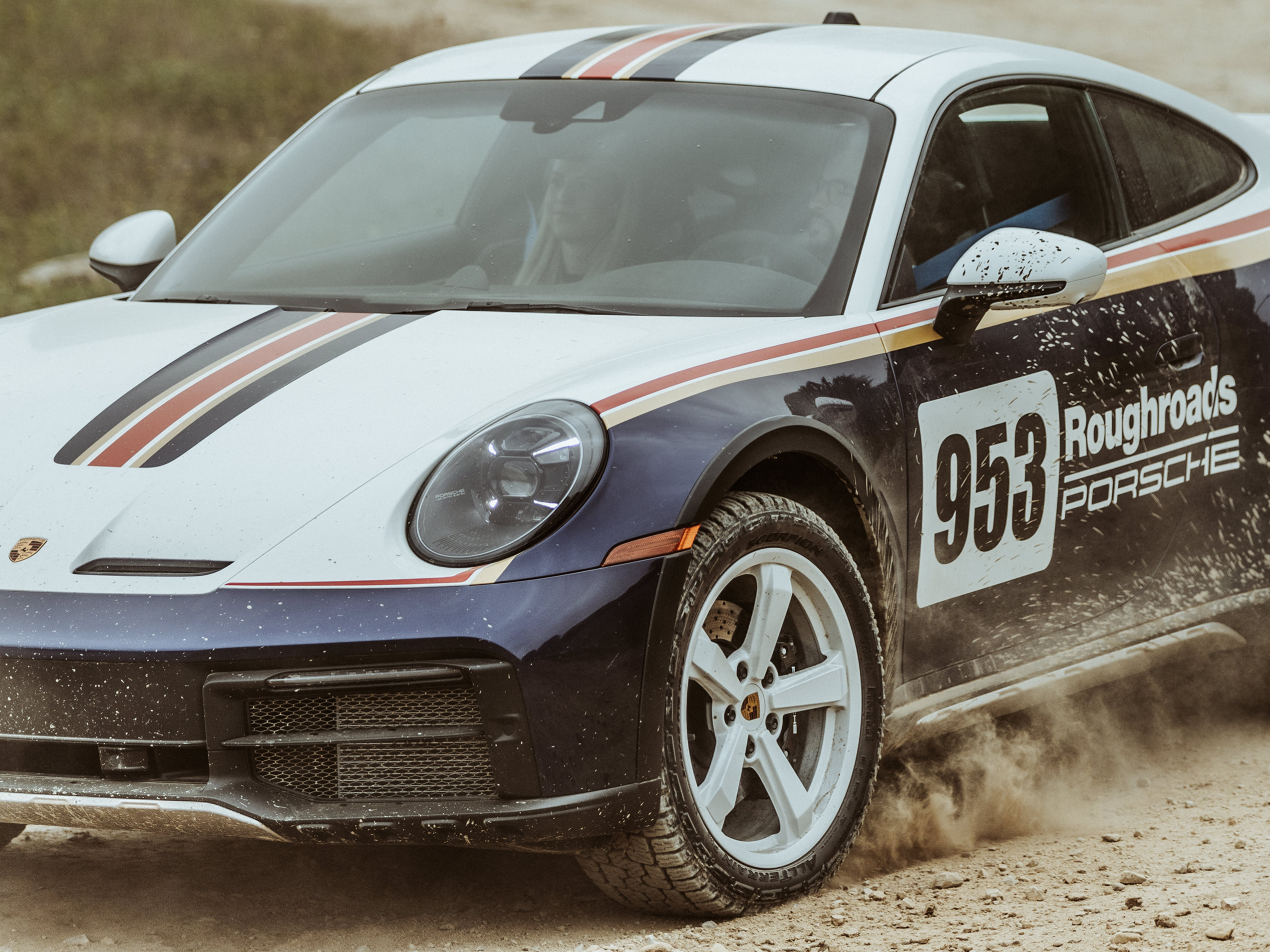 Ein Porsche in verschiedenen Lackierungen fährt durch Outback und wirbelt Sand auf.