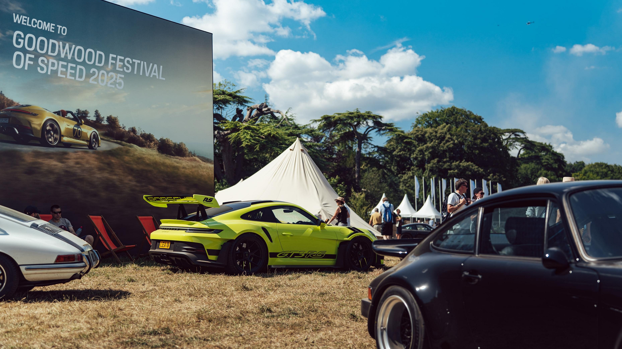 Festivalgelände vom Goodwood Festival mit Posterwand und mehreren Porsche Modellen.