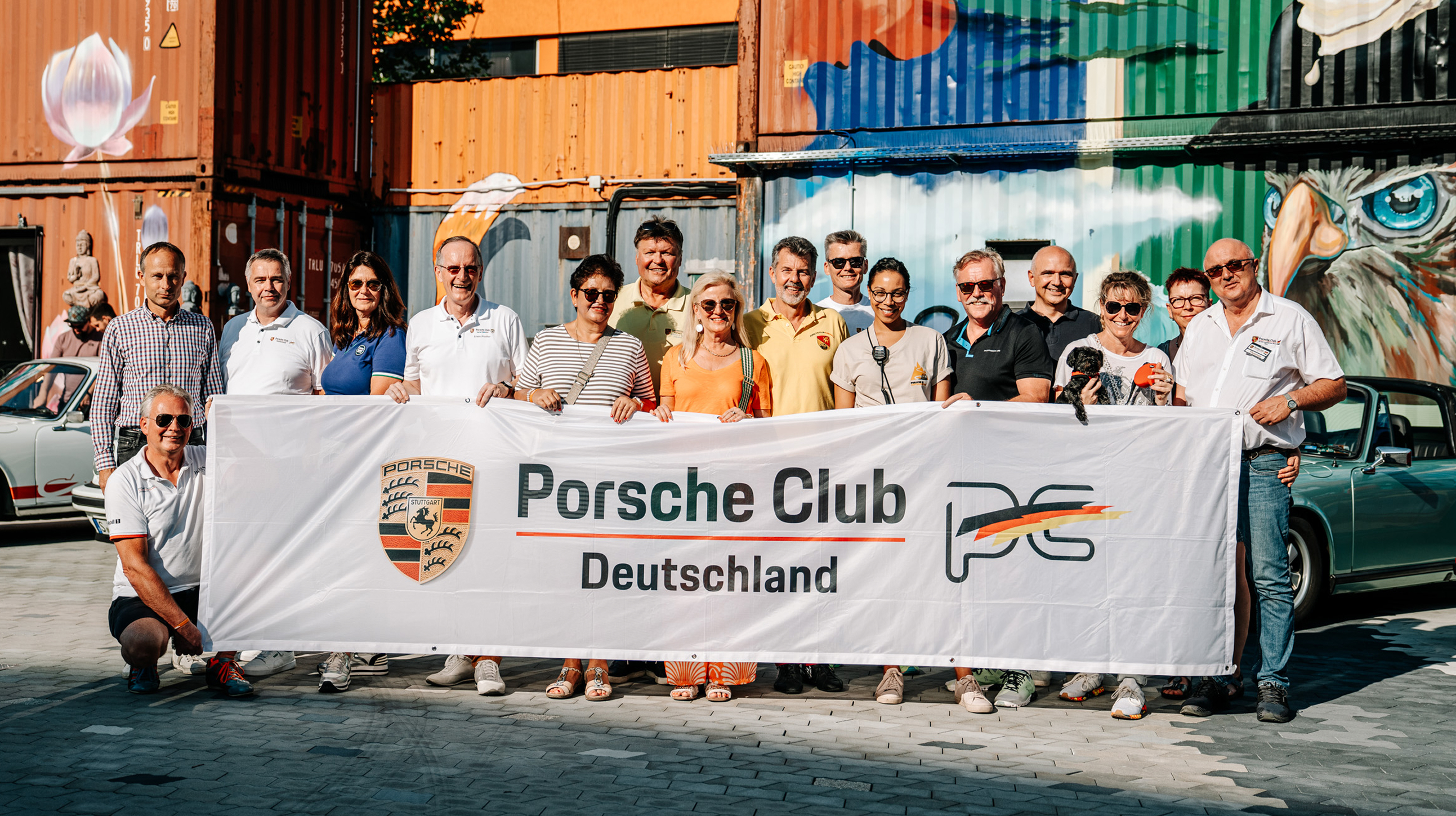 Mehrere Menschen halten ein Banner hoch mit der Aufschrift "Porsche Club Deutschland". Im Hintergrund bemalte Container.