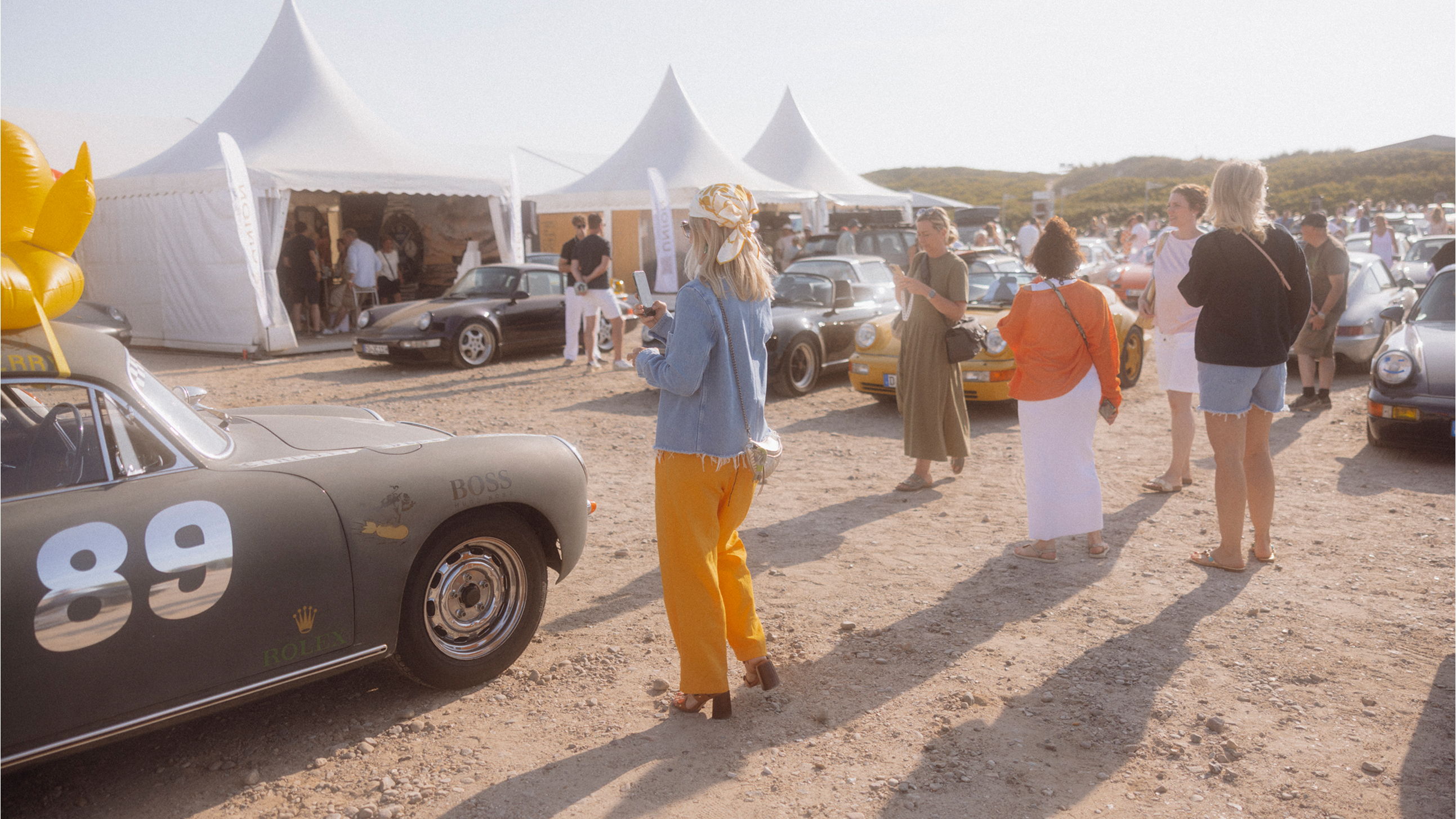 Mehrere Menschen stehen zwischen Zelten und Porsche Fahrzeugen in Sylt am Strand beim Petro Surf Festival.