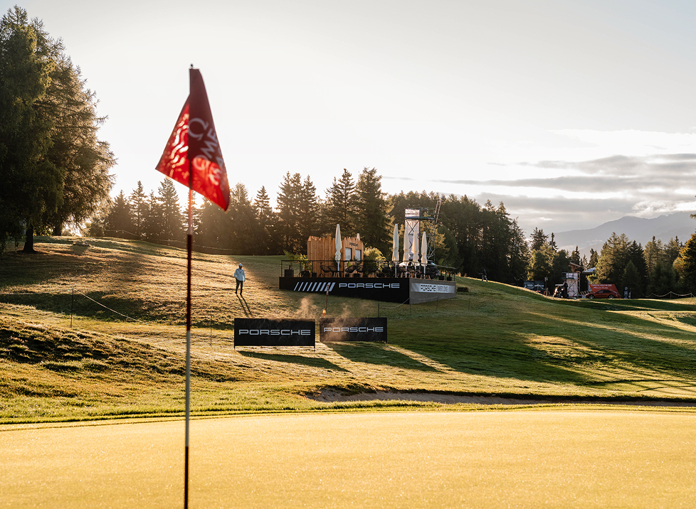 Golf Kurs in Andermatt mit Aufbauten von Porsche bei Sonnenaufgang. Im Vordergrund ein Fahne.
