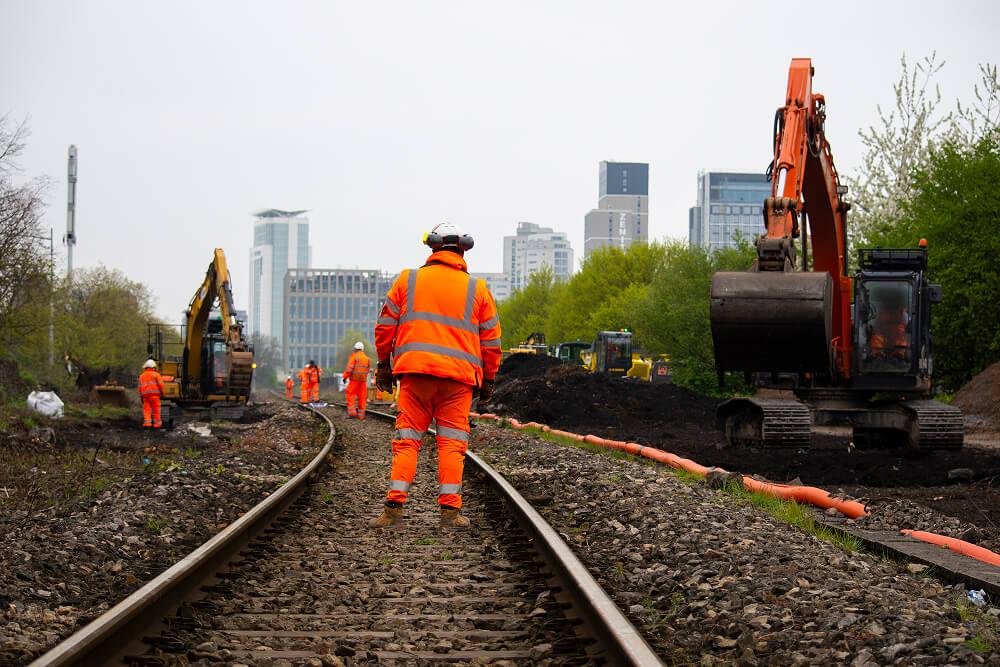 Construction workers in orange high-visibility clothing on a railway track with excavators, against a cityscape background with trees and buildings.