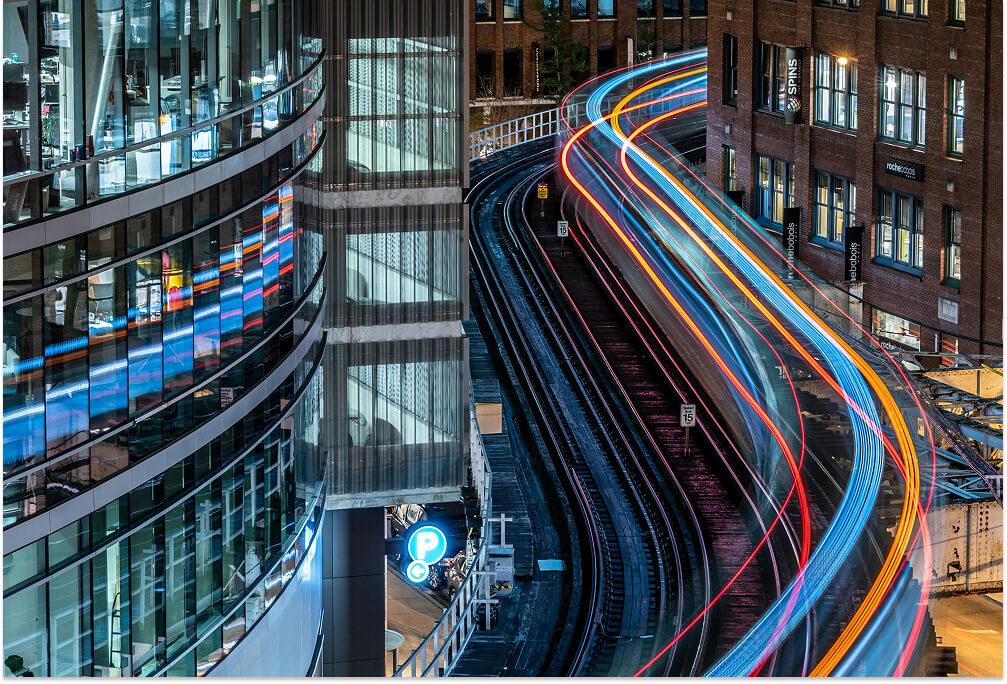 Night view of a city rail track with colorful light trails from passing trains, flanked by glass buildings, reflecting vibrant lights.