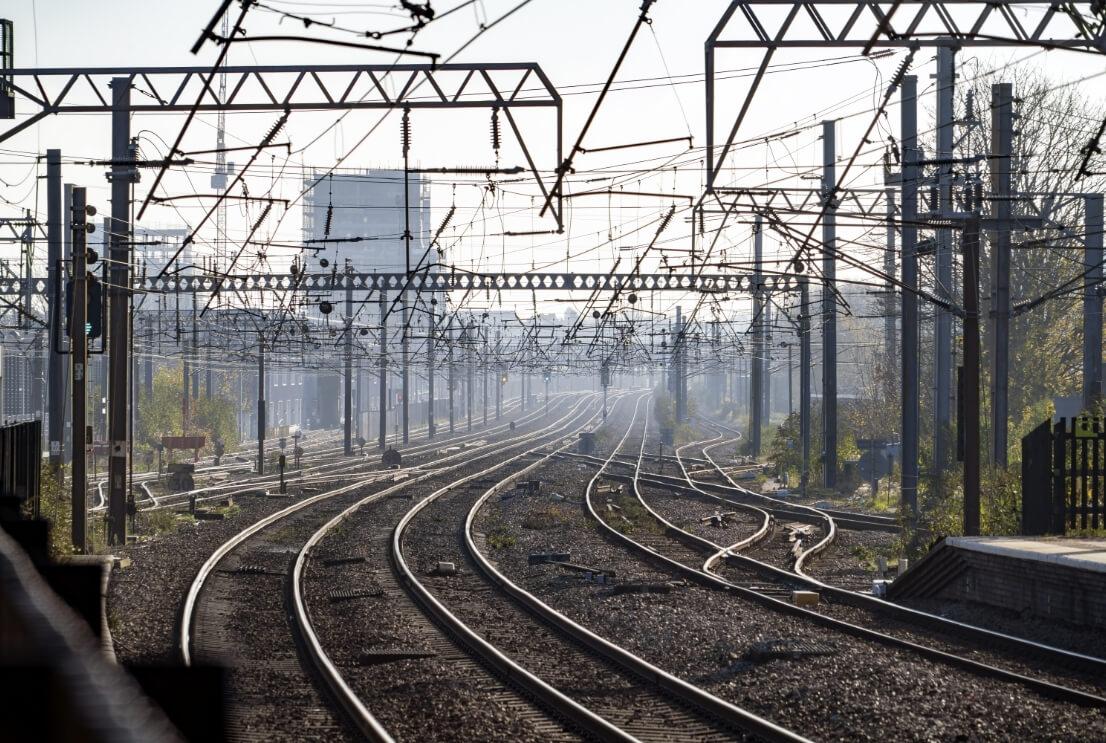 Railway tracks with overhead power lines curve into the distance, surrounded by industrial structures and a hazy sky.