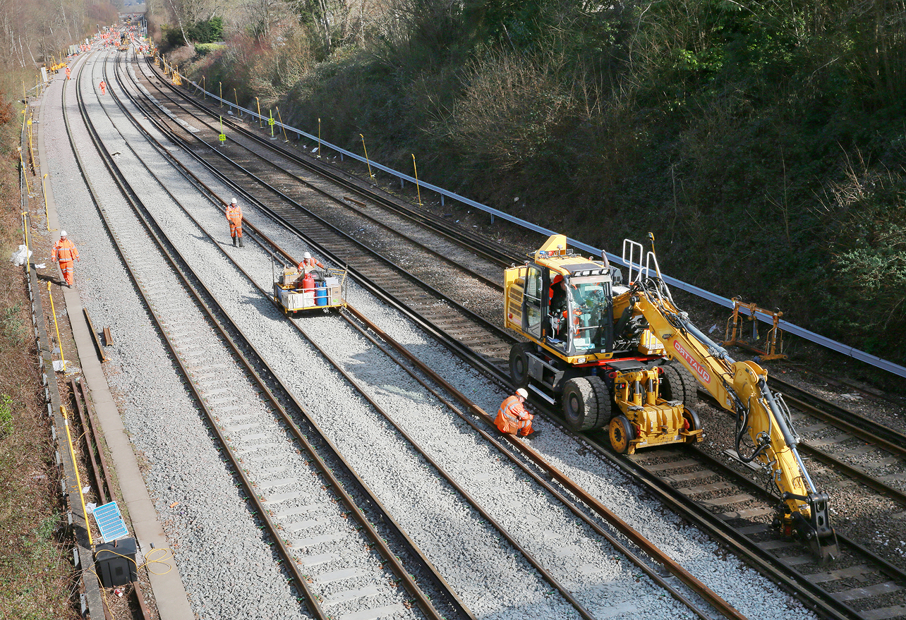 Railway maintenance scene with workers in orange uniforms and machinery on tracks, surrounded by trees and gravel, under a clear sky.