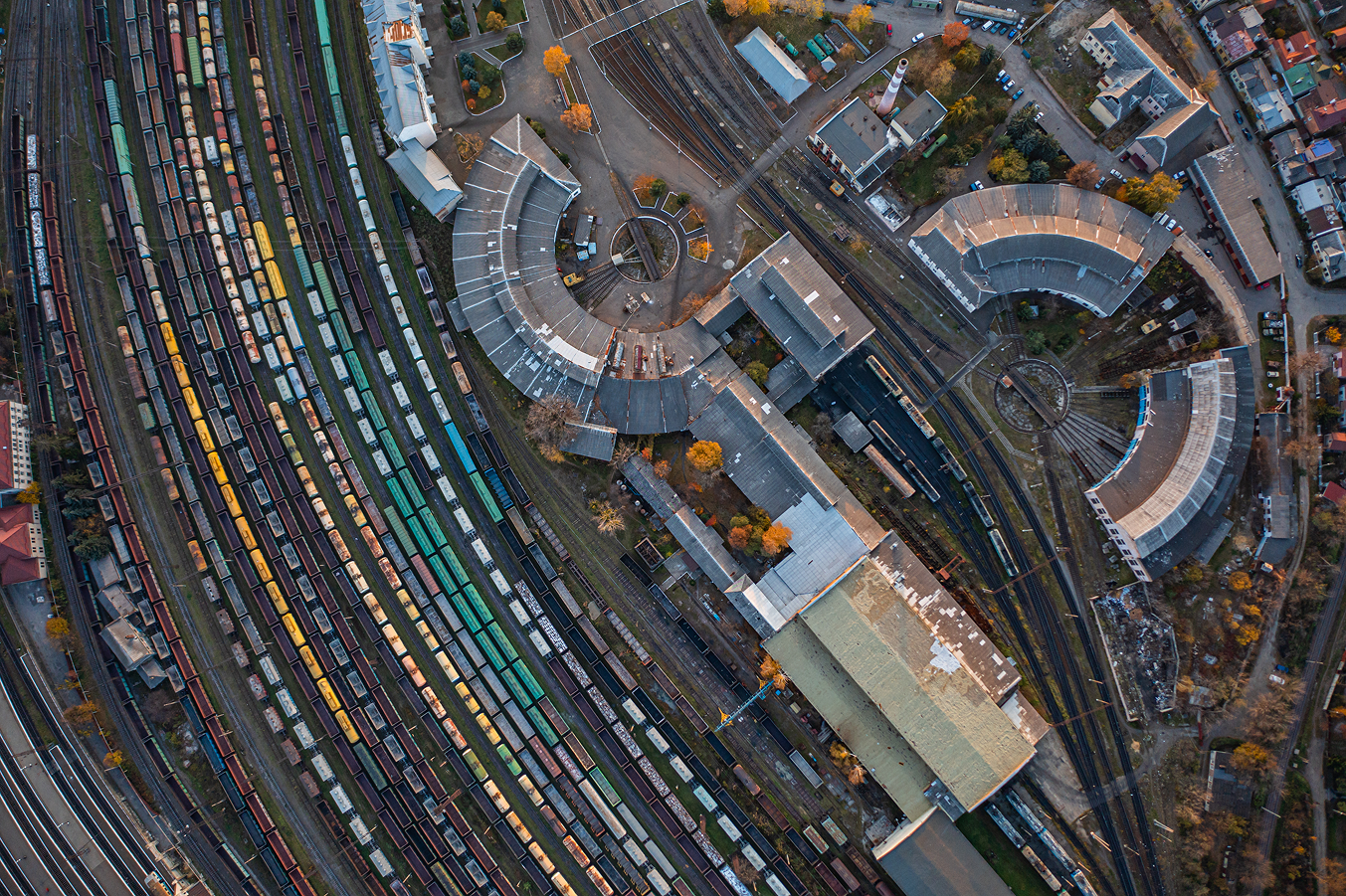 Aerial view of a railway yard with numerous colorful train cars on curved tracks, surrounded by industrial buildings and trees with autumn foliage.