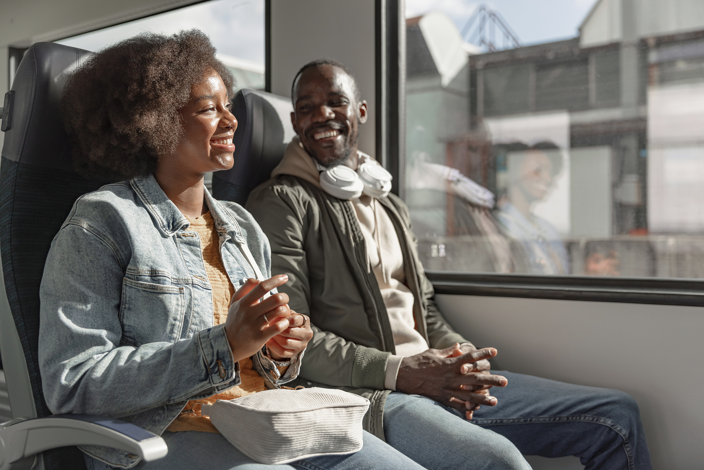 Two people sit in a train, smiling and talking. One wears a denim jacket, and the other has headphones around their neck. Sunlight streams in.