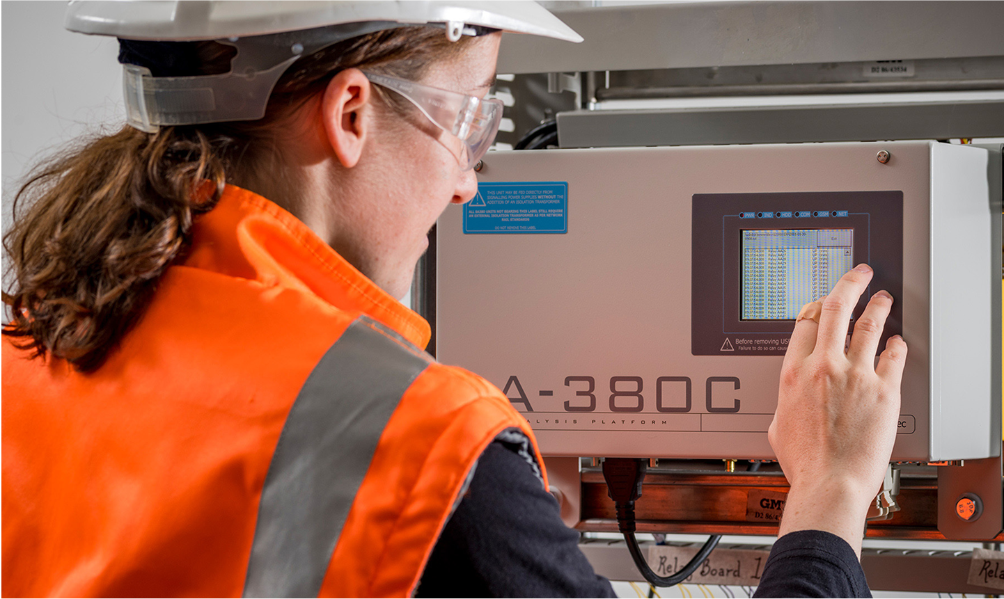 Person in safety gear operates a touchscreen on industrial equipment, wearing an orange vest, hard hat, and safety glasses.