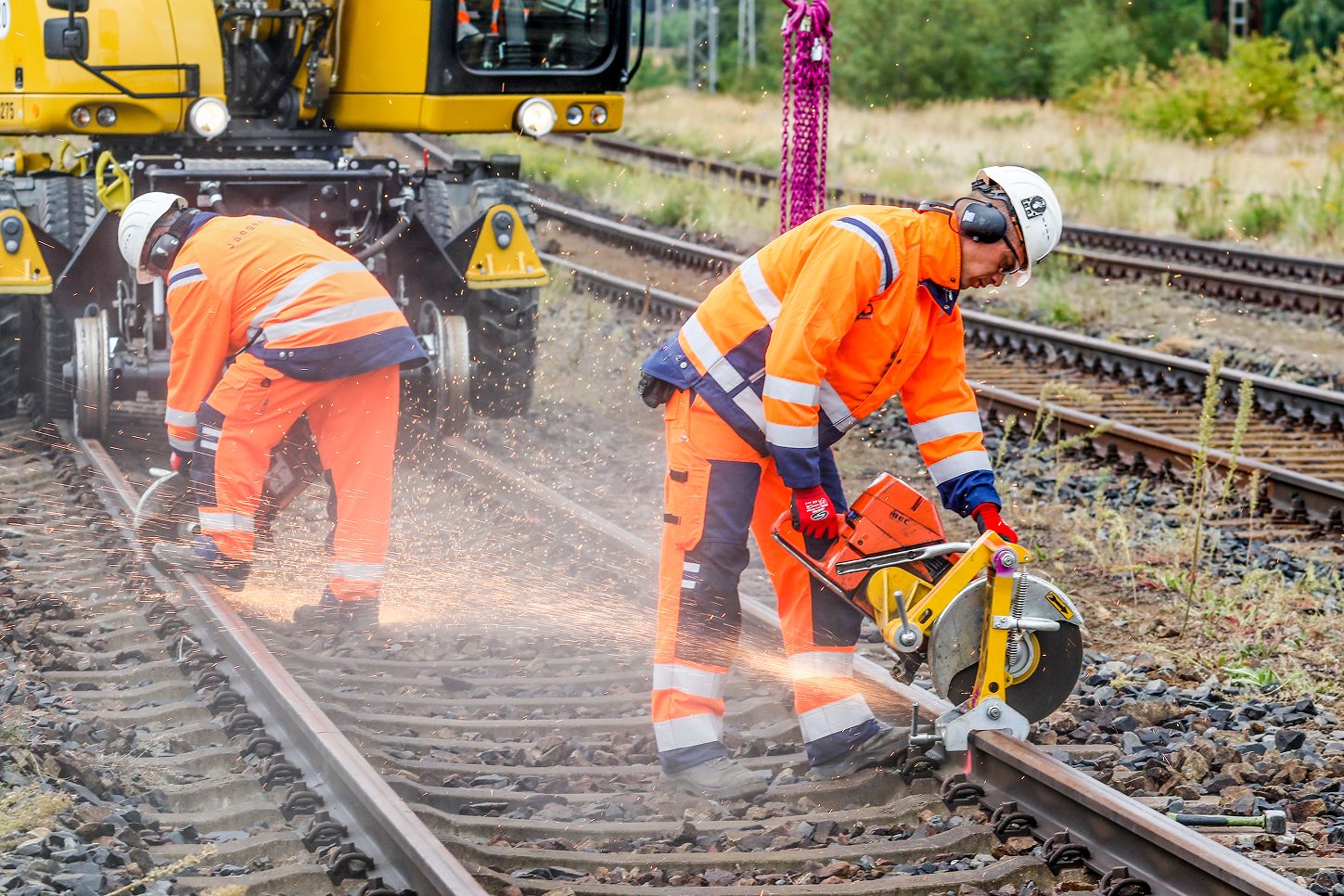 Workers in orange safety gear cutting railroad tracks with a saw, surrounded by equipment and greenery in the background.