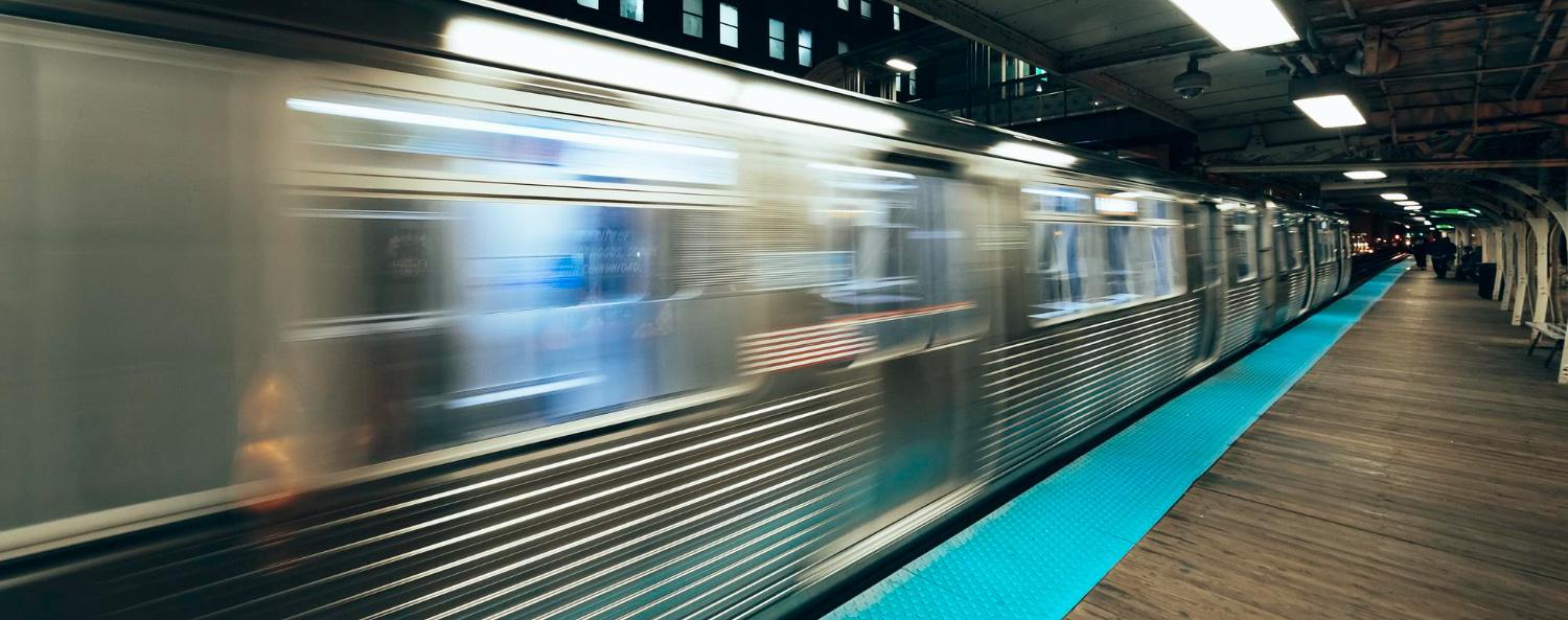 A fast-moving subway train blurs past an elevated platform with blue safety markings, under a roof with exposed beams and lights.
