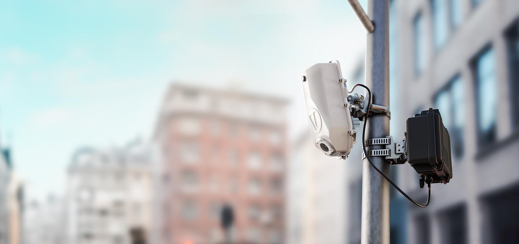 A white security camera mounted on a pole, overlooking a blurred urban street with buildings in the background.