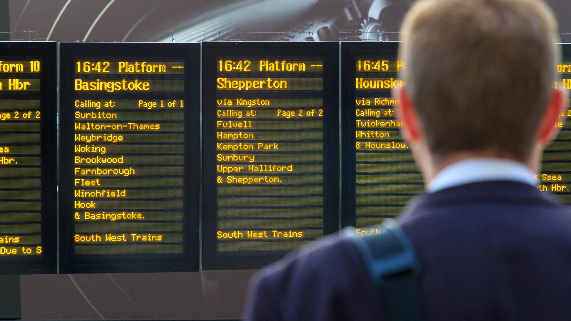 A person stands in front of a train schedule board displaying destinations like Basingstoke, Shepperton, and Hounslow with departure times.