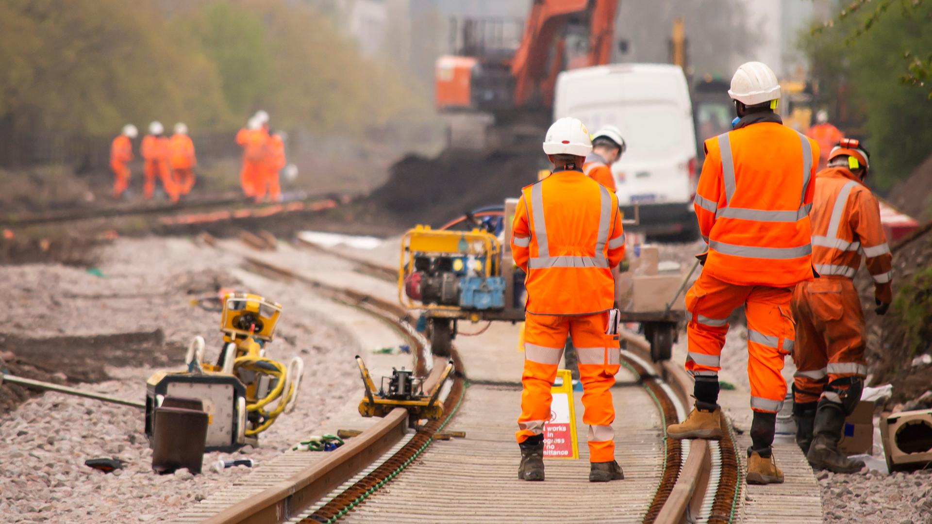 Construction workers in orange safety gear work on railway tracks, with machinery and vehicles in the background.