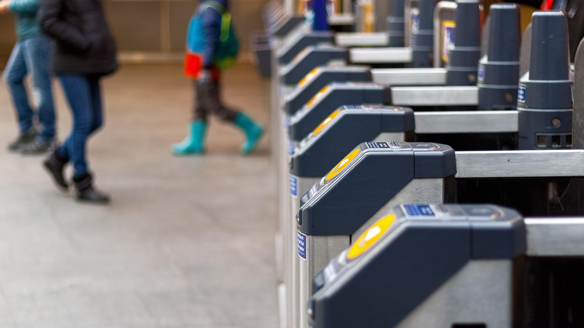 Close-up of electronic ticket barriers at a transit station entrance, with blurred people walking in the background.