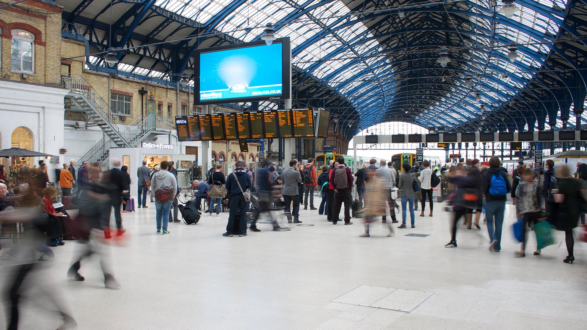 Busy train station interior with people walking, a large digital screen, and a departure board under a high, arched ceiling.