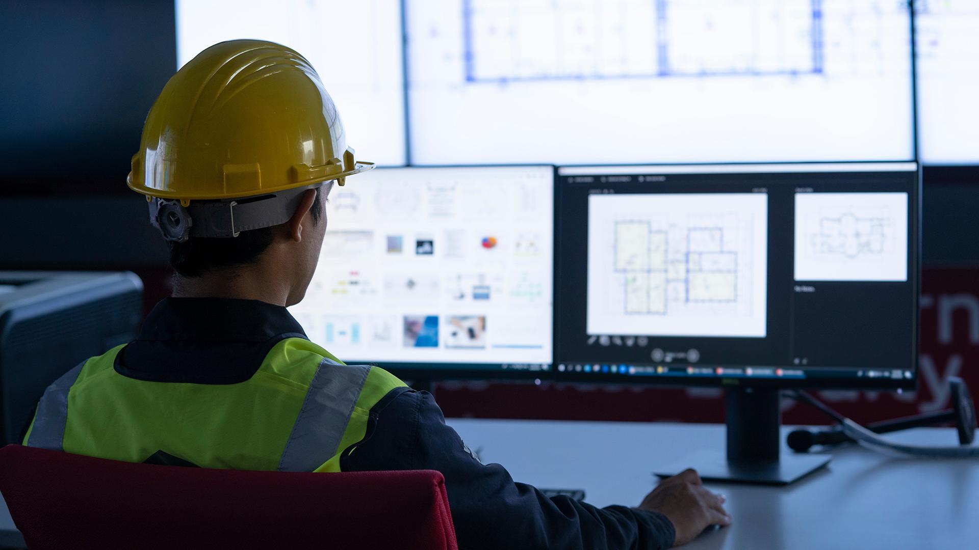 Person in a yellow hard hat and reflective vest working on architectural designs at a dual-monitor setup in an office.