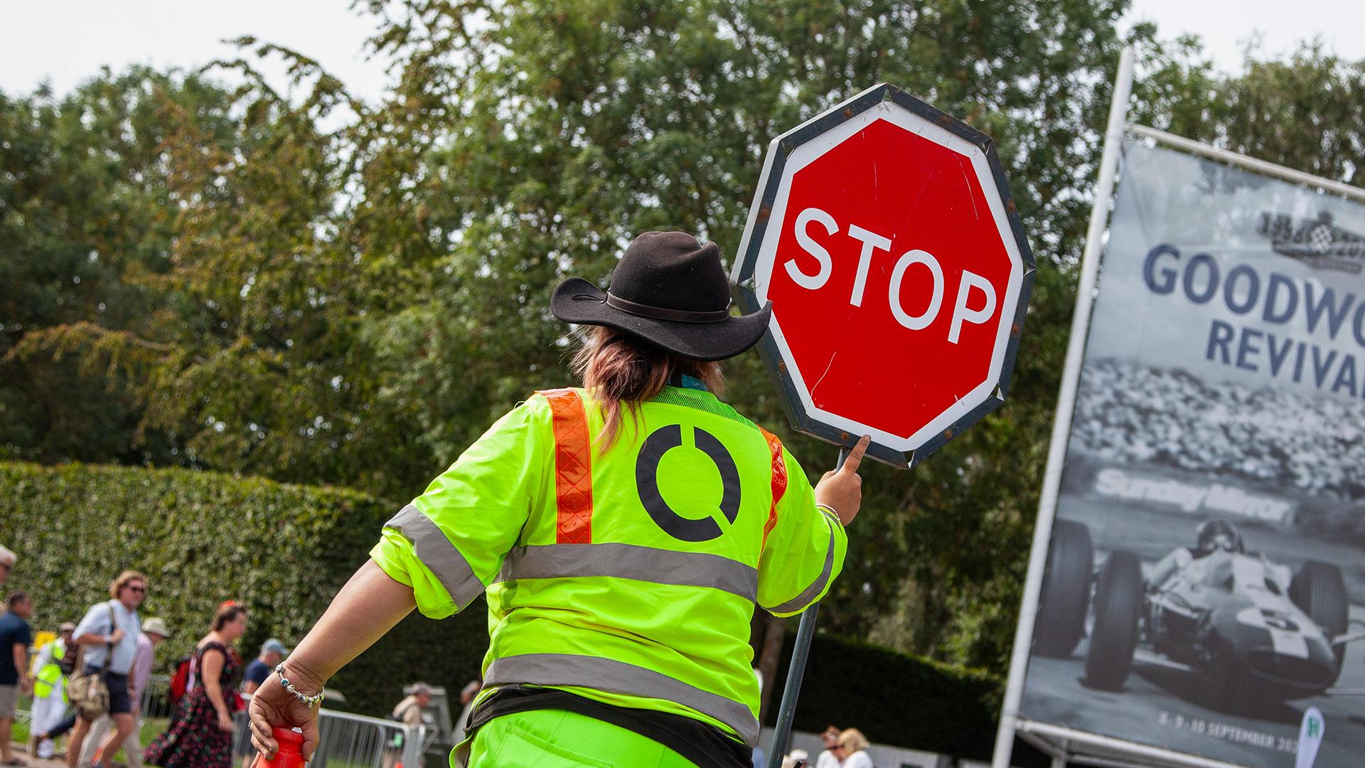 A person in a neon safety vest and black hat holds a stop sign at an outdoor event, with trees and a large poster in the background.